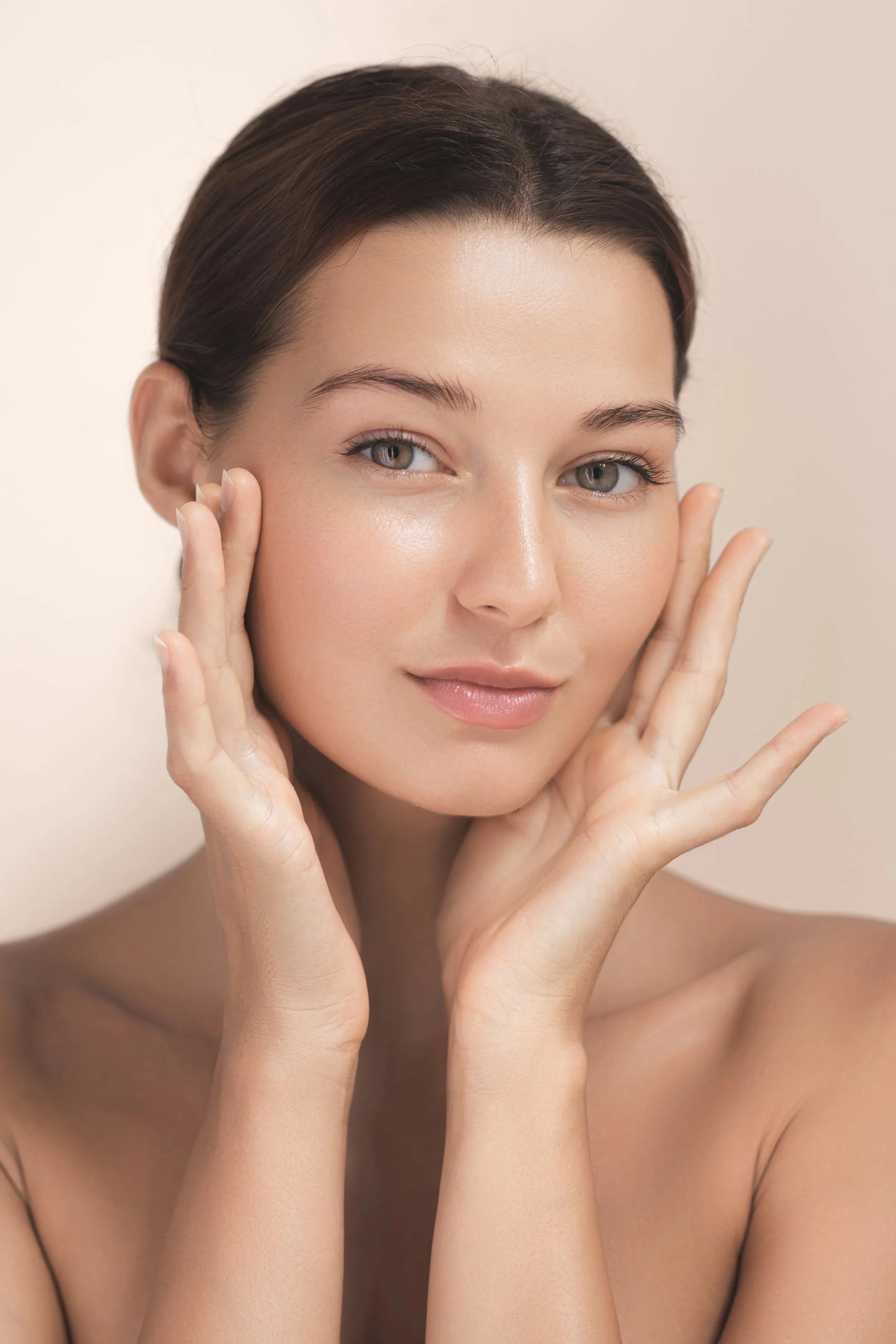 Close-up of a young woman with clear skin, gently touching her face with both hands, smiling softly, against a neutral background.
