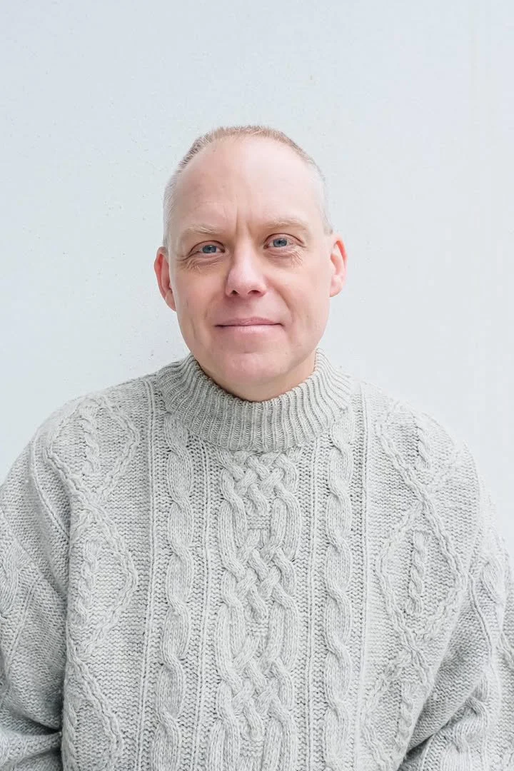 A man with short blond hair and blue eyes wearing a light gray cable-knit turtleneck sweater, standing against a plain light-colored wall.
