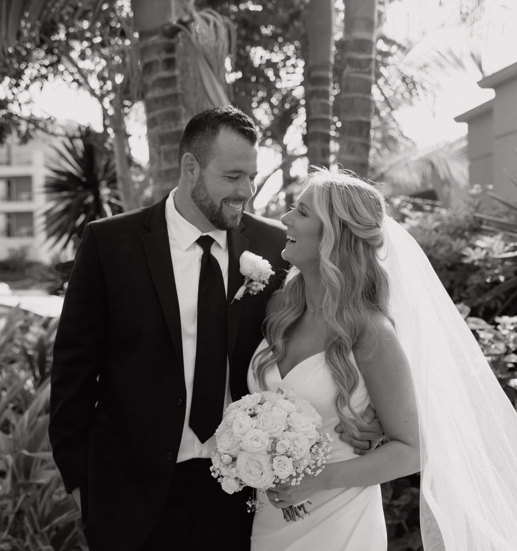 Black and white photo of a bride and groom on their wedding day outside, smiling and looking at each other. The bride has long wavy hair and is holding a bouquet of roses, wearing a strapless wedding dress and veil. The groom has short hair and a beard, wearing a black suit with a white shirt, black tie, and a boutonniere. Palm trees and plants are in the background.