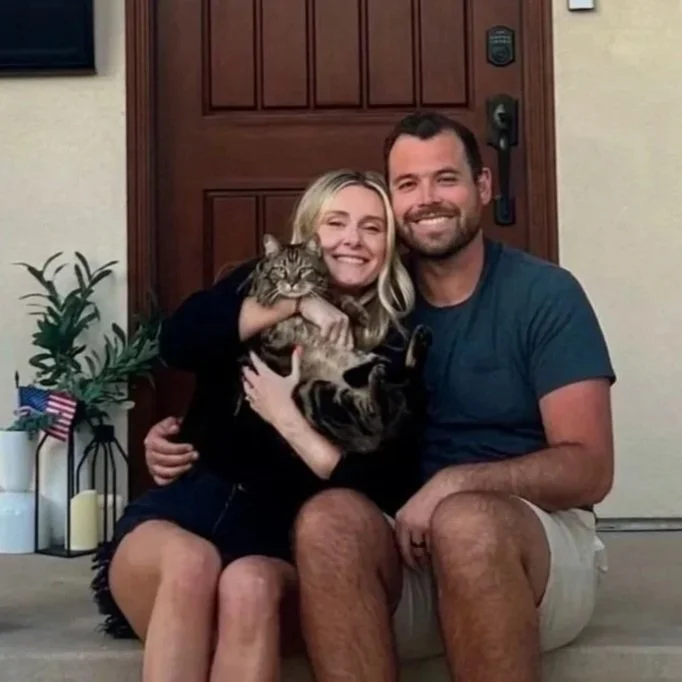 Happy couple sitting on front porch steps, holding a tabby cat, smiling at the camera, with house door and potted plants in the background.
