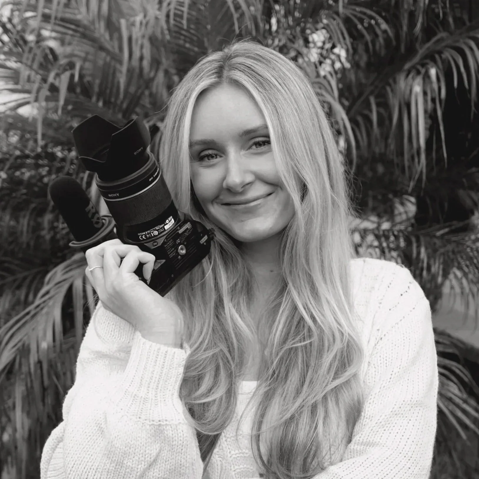 Black and white photo of a woman with long, wavy hair smiling and holding a professional camera with a large lens and lens hood, standing in front of lush foliage.