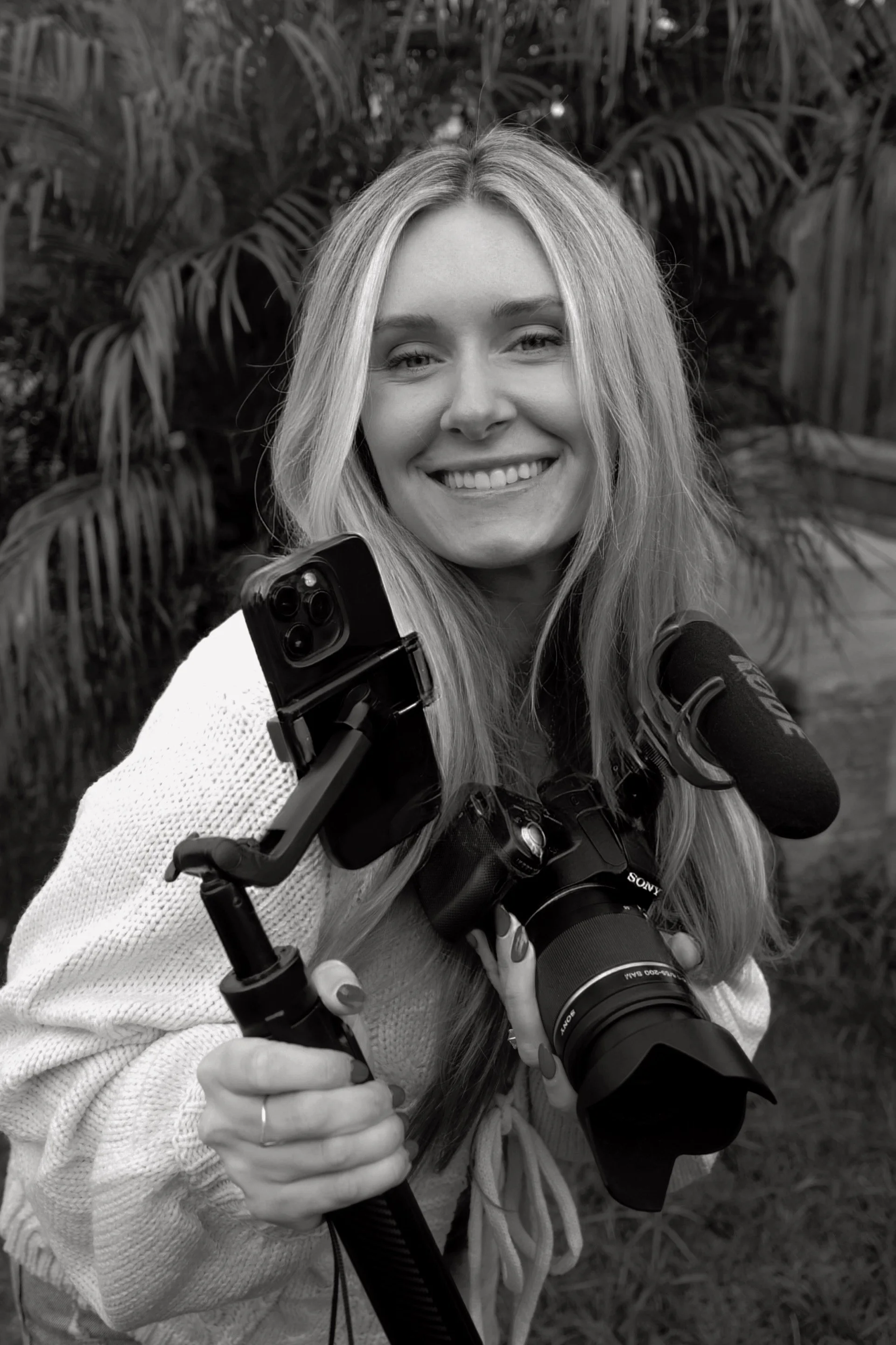 Black and white photo of a smiling woman holding a camera on a tripod with a smartphone mounted on it, outdoors with palm trees in the background.