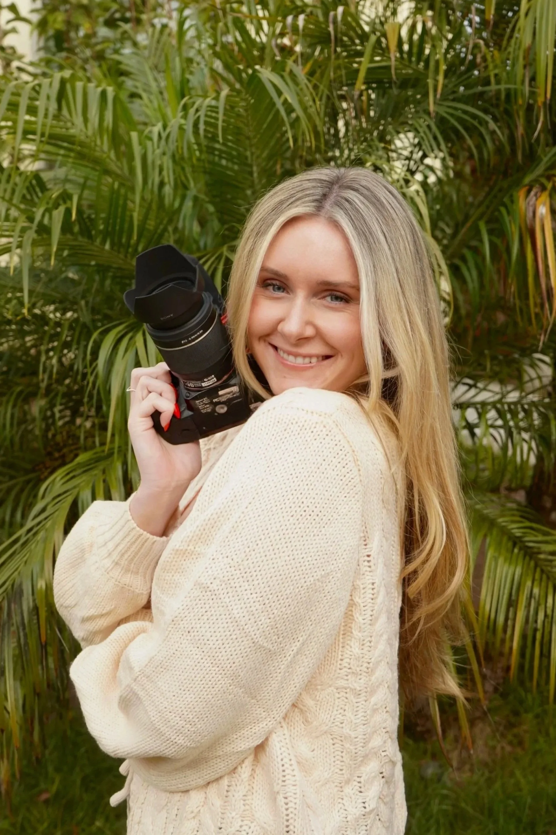 A young woman with long blonde hair smiling and holding a camera, standing outdoors in front of green palm leaves.