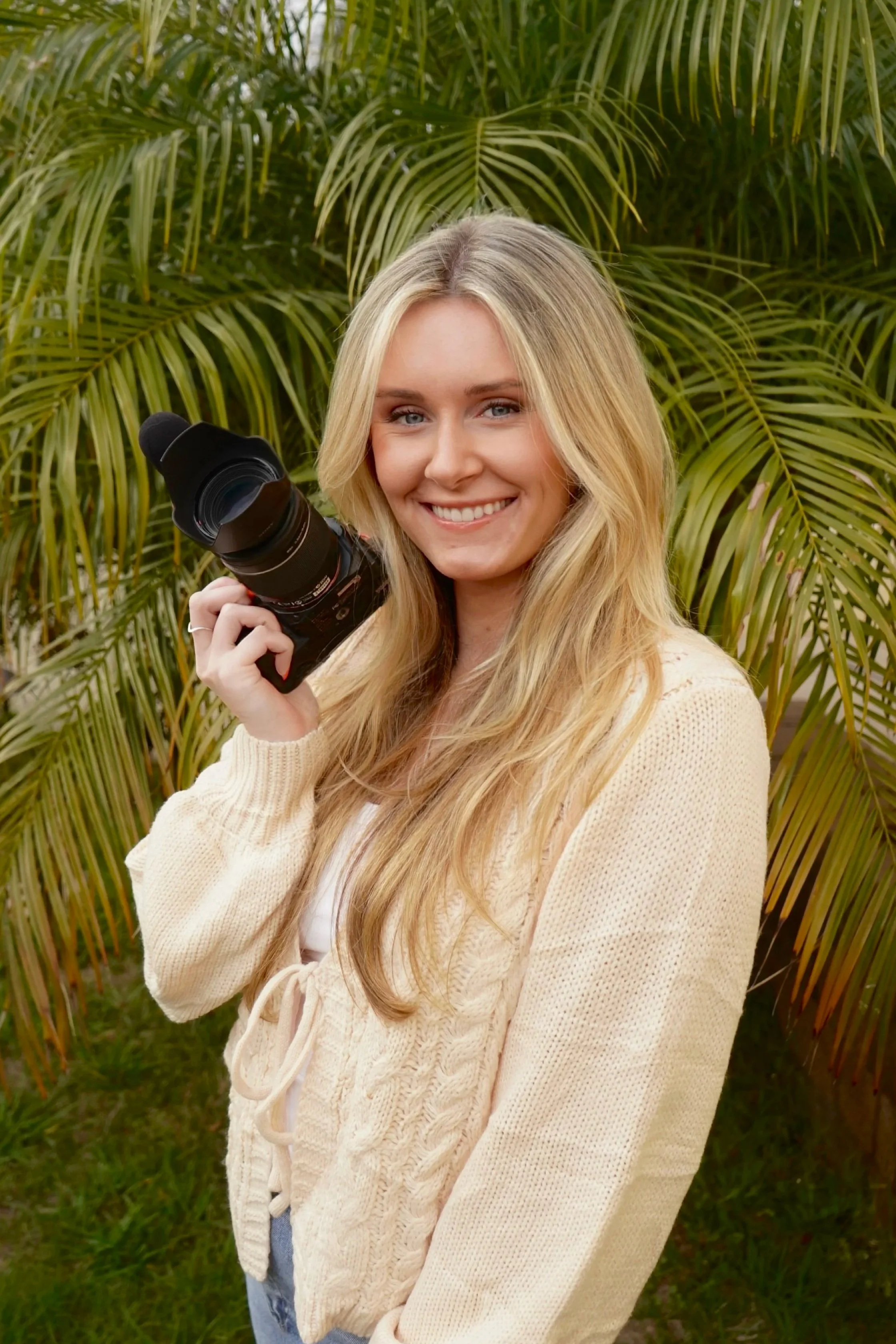 A young woman with long blonde hair smiling and holding a camera in front of green palm leaves.