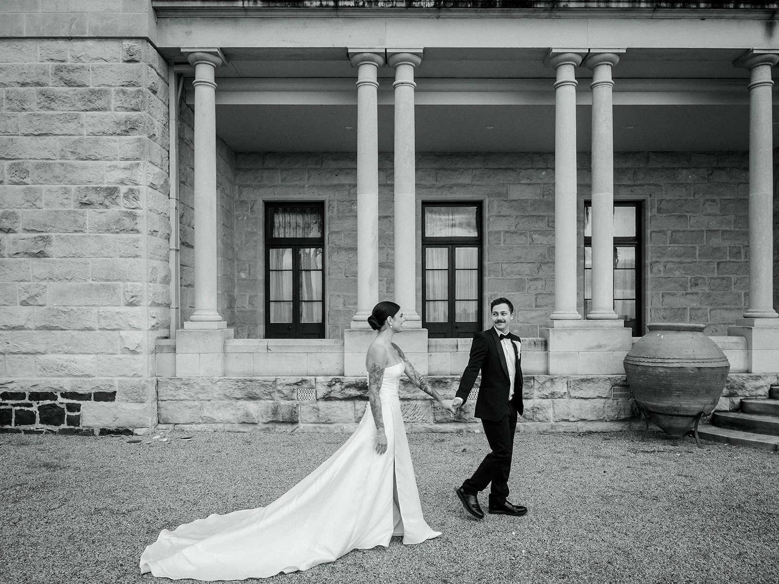 Black and white portrait of Cherry and Nick walking hand in hand outside Jimbour House colonnade — Sean Reefman Photography