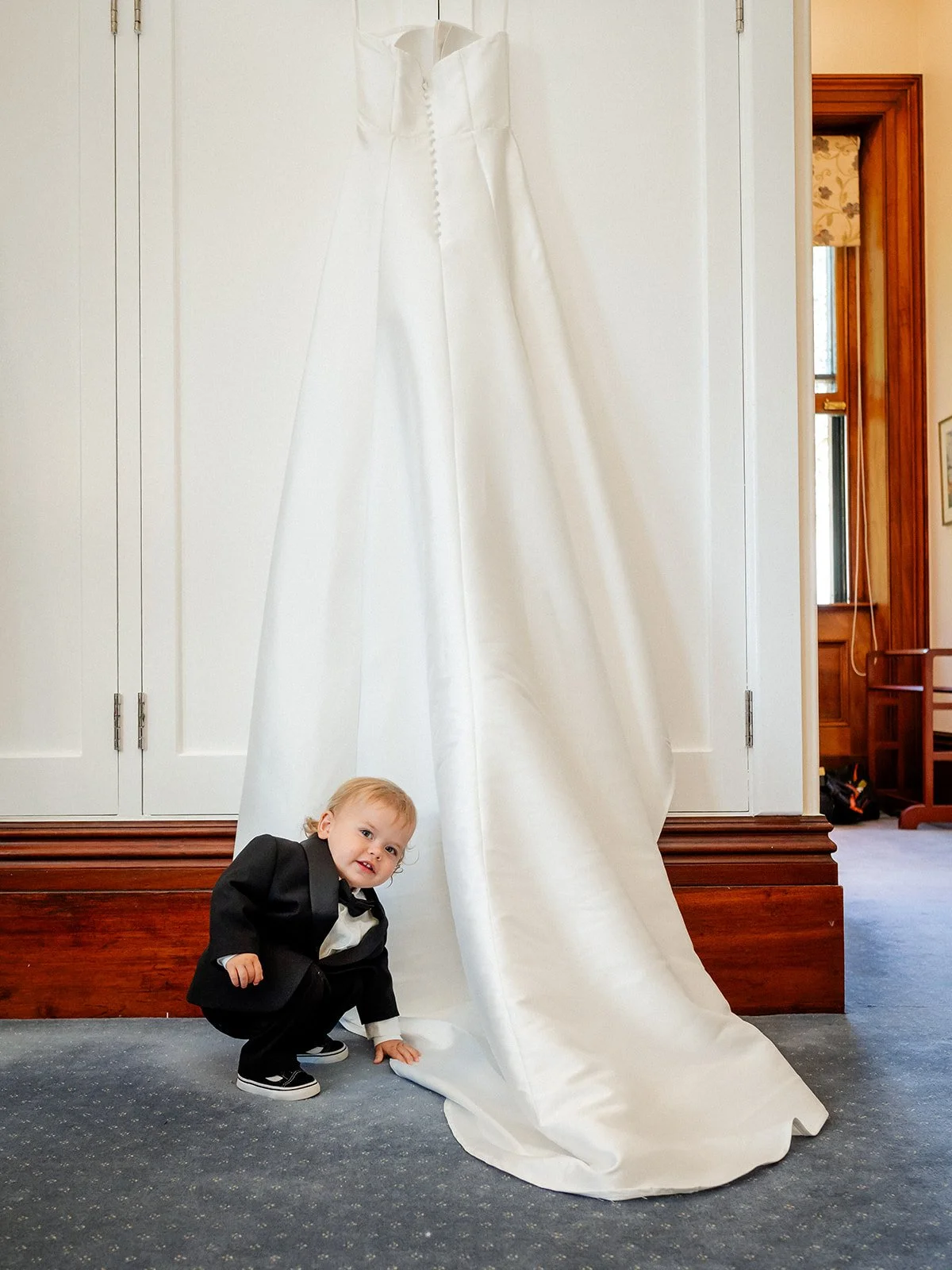 Wedding dress hanging inside Jimbour House with baby in tuxedo crouching beneath — Sean Reefman Photography