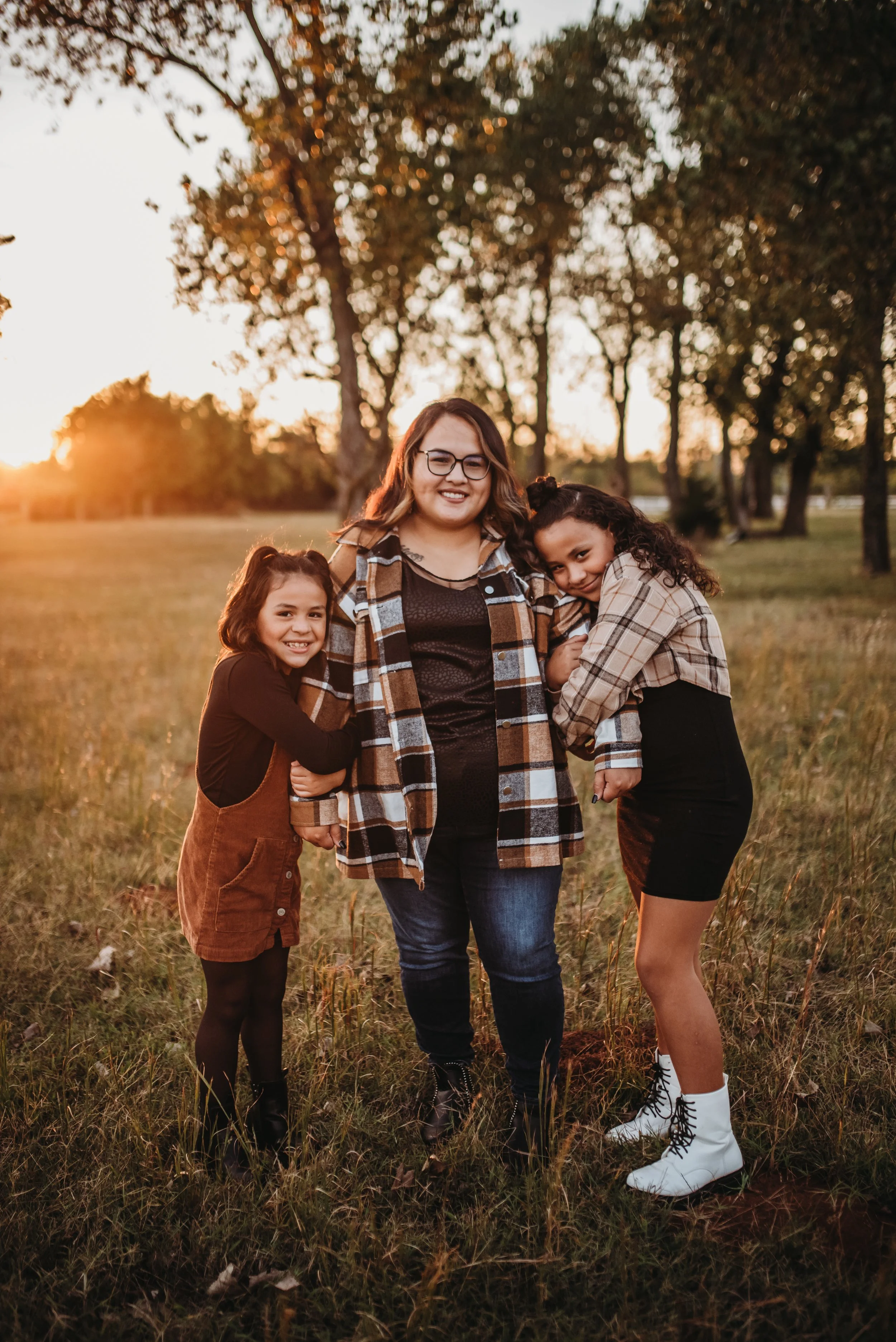 A woman in a plaid jacket stands in a field with two young girls, one in a brown dress and the other in a plaid shirt, under a sunset sky with trees in the background.