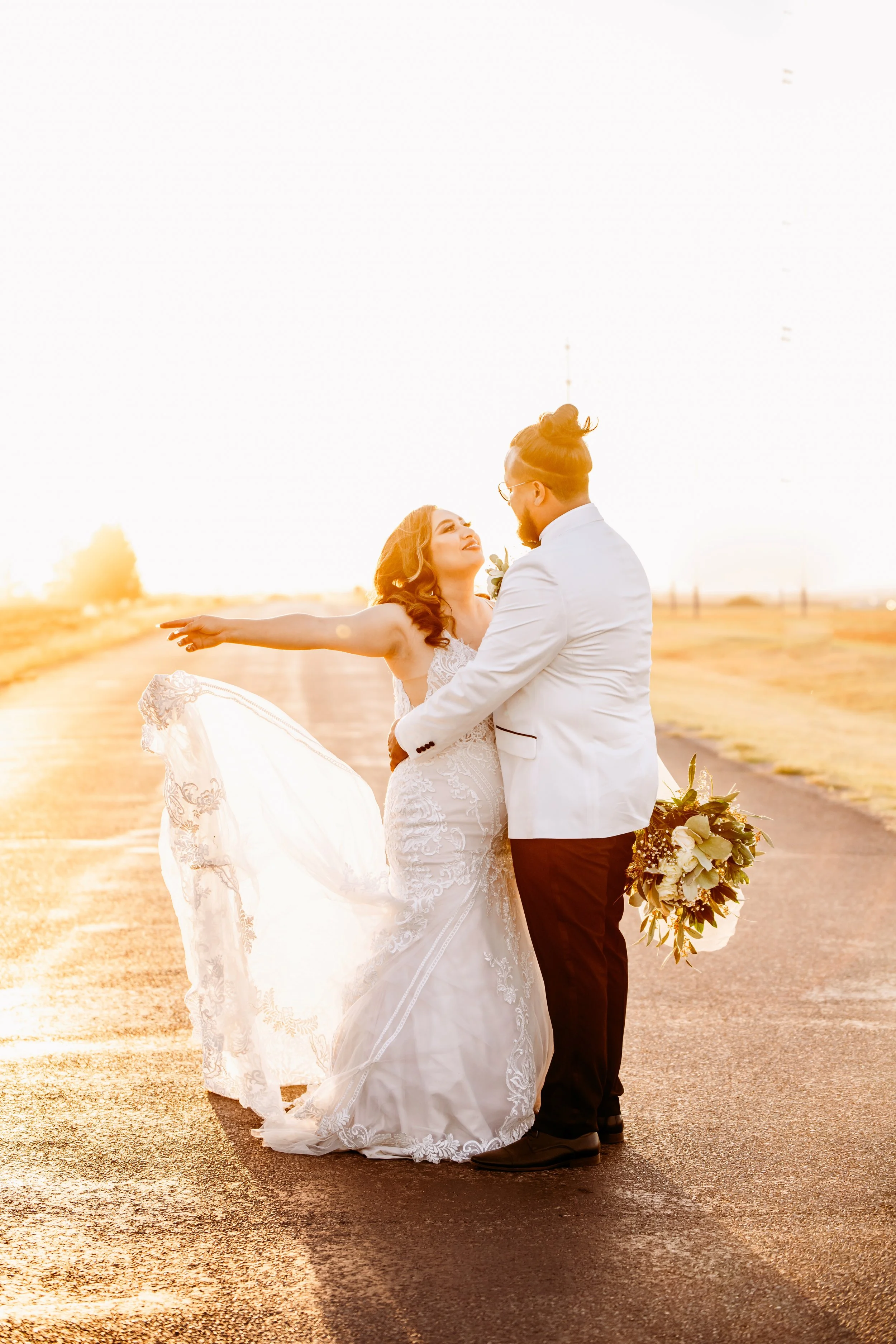 Bride in lace wedding gown and groom in white suit embracing, holding bouquet, on sunlit road.