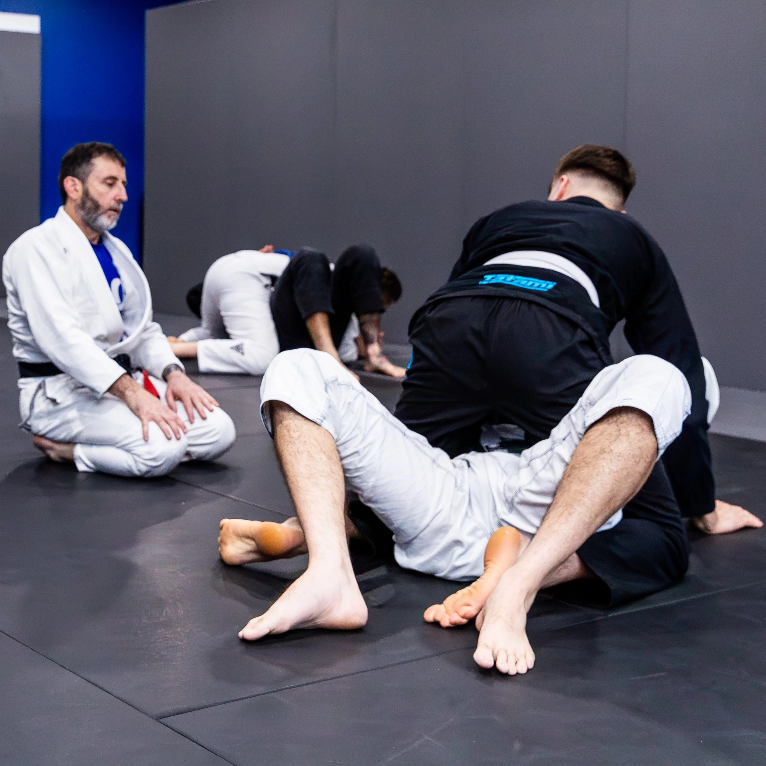 Coach Trent is kneeling in a white gi next to two students. Trent is talking the students through a technique. One student is kneeling on top of the other, using his hips to keep his partner on the ground. The student on top is wearing a black gi.