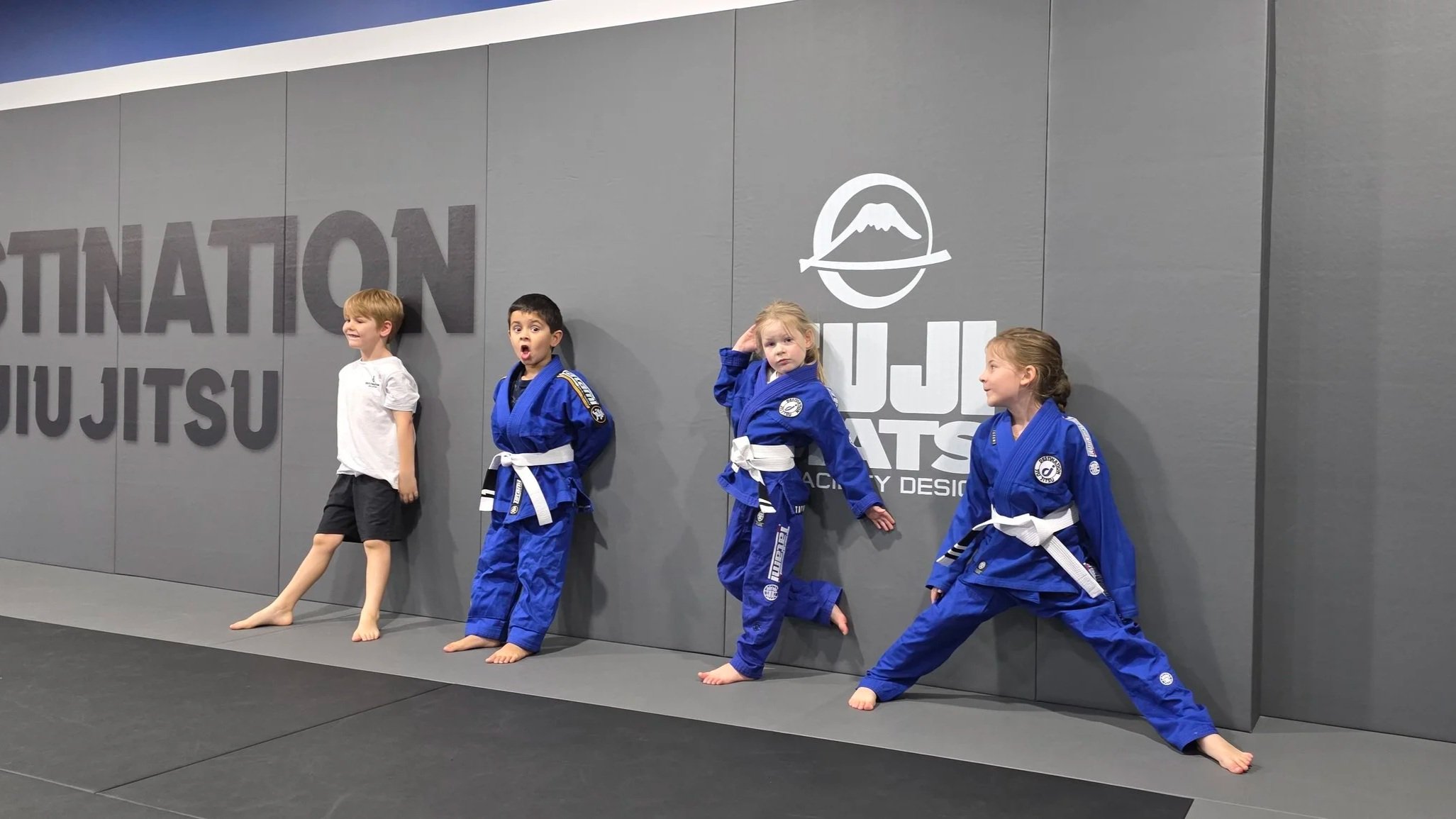 Four young students are standing in different poses against the wall. One student is wearing a white t-shirt and black shorts. The other three students are wearing blue gis with white belts.