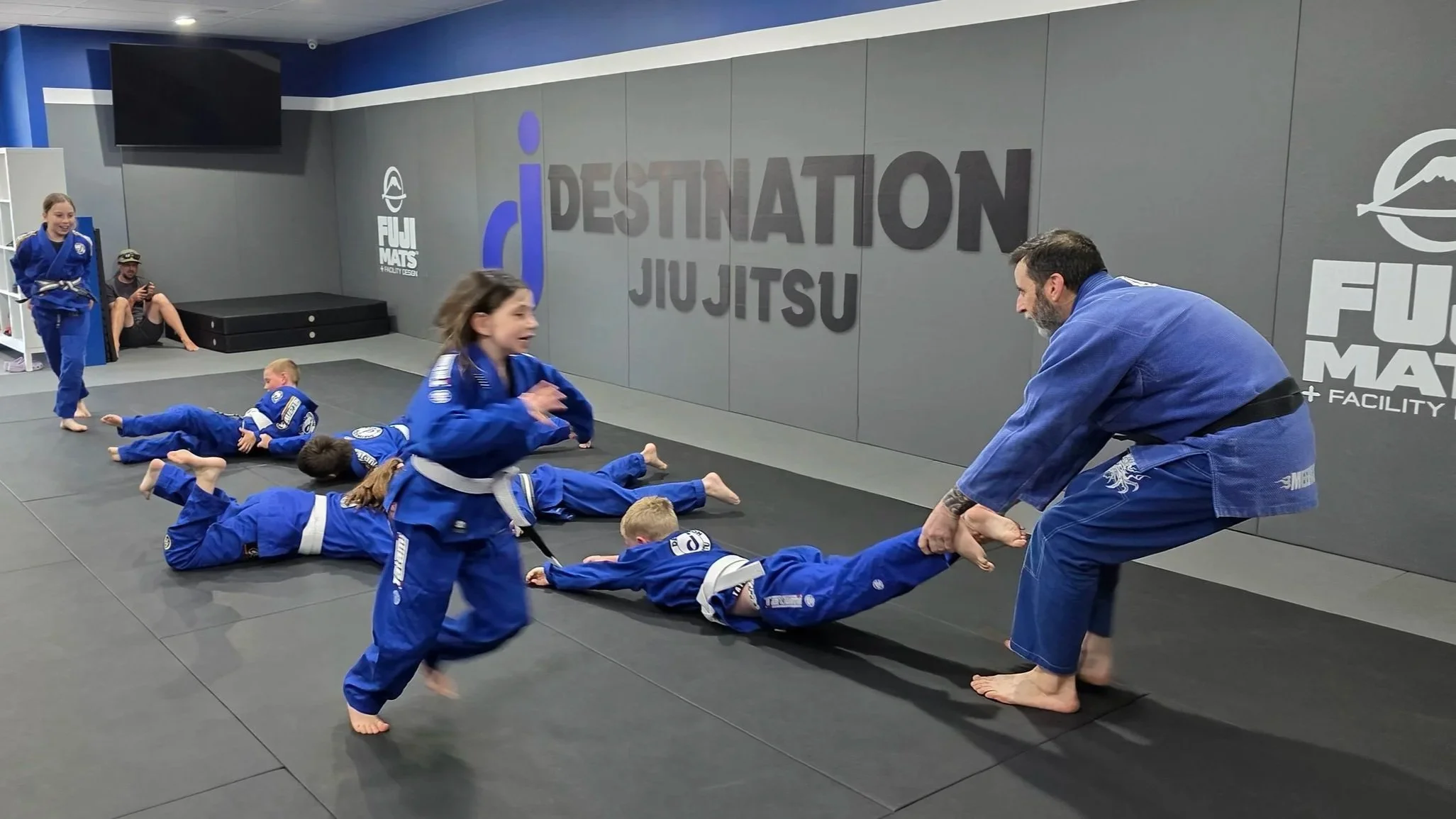 This image shows a kids class playing a jiu jitsu game. FIve students are laying on the ground, with the coach holding the ankles of one student. Two students are running around the others. All students are wearing blue gis. 