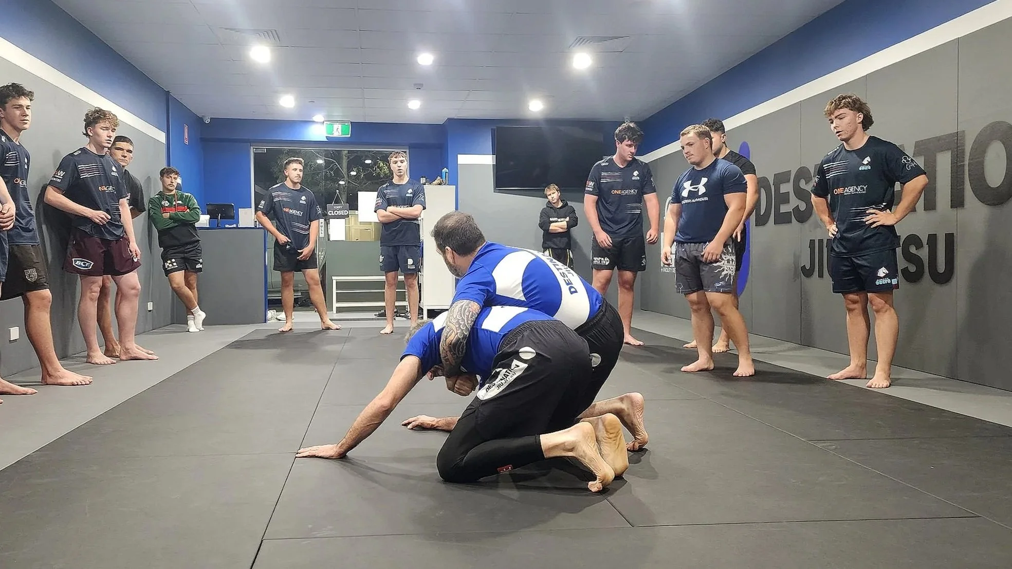 A group of young men dressed in t-shorts and shorts are standing in a semi circle watching the coach and an assistant demonstrate a BJJ technique while on their hands and knees. The coach and assistant are dressed in blue and white rashguards and bla