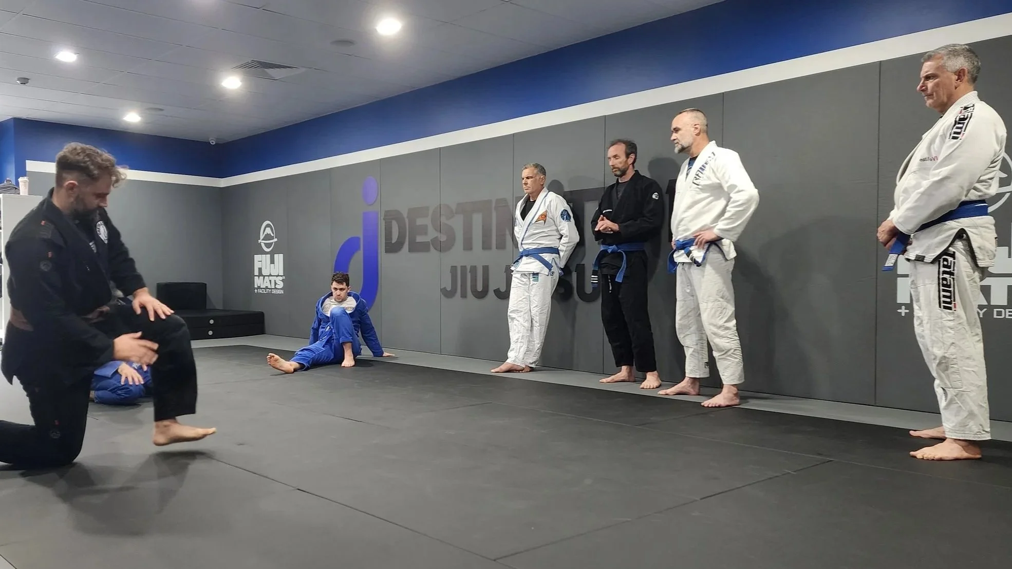 A group of men in BJJ kimonos are watching a demonstration of a BJJ technique. A man is on one knee in the foreground. 