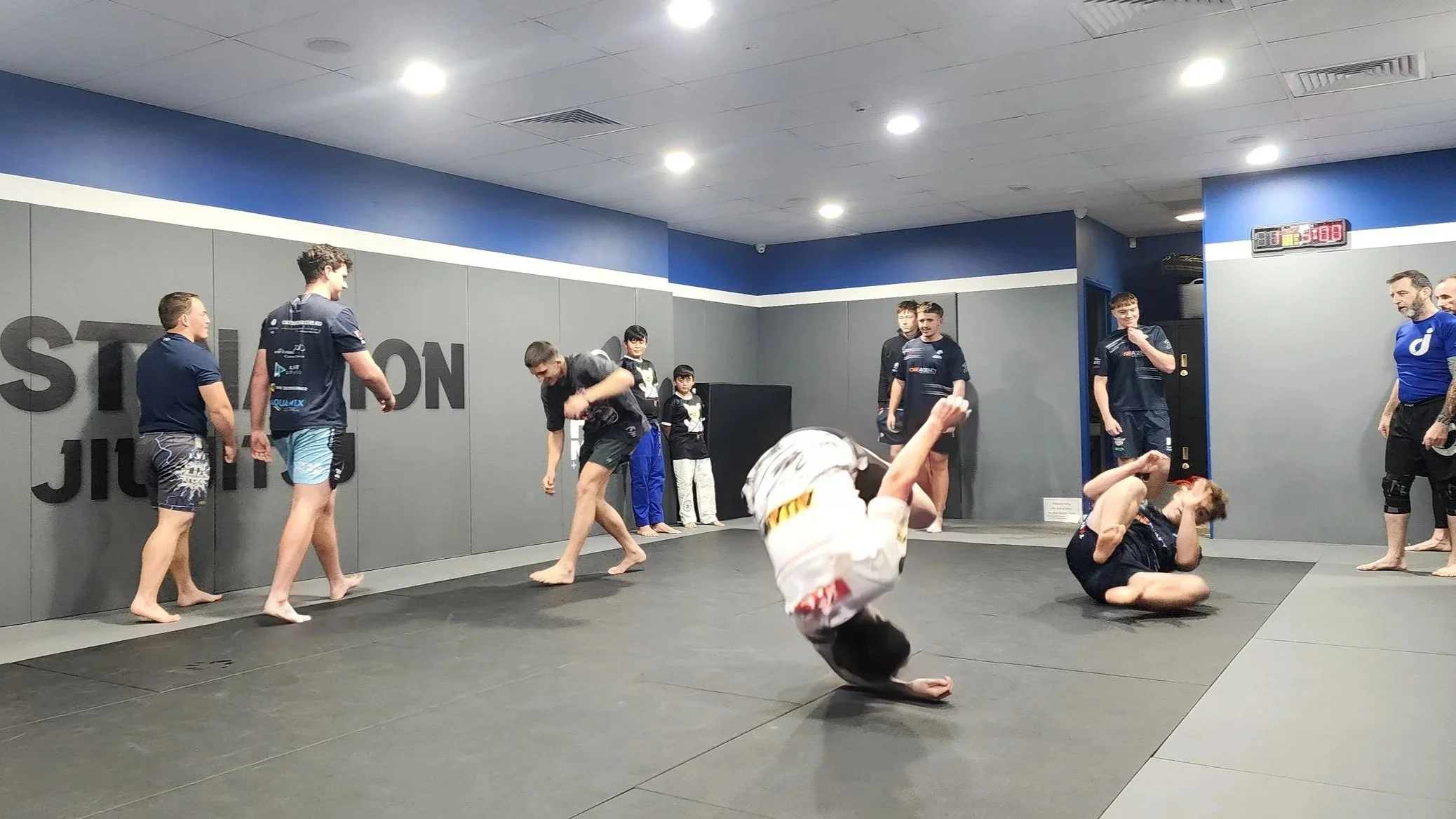 A group of young men are completing a warmup exercise of forward rolls down the length of the matted space. Two are actively rolling, while the others are walking in the background to start the next warm up exercise. The group are dressed in shorts a