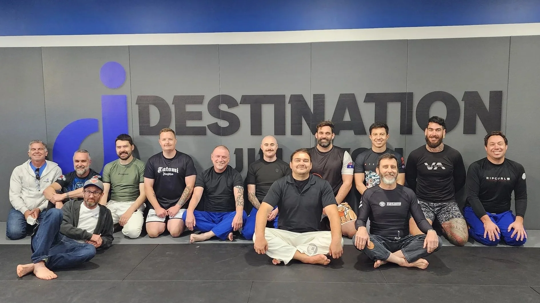 A group shot of participants after a self-defence seminar. Participants are all male, and are wearing rashguards and loose pants, and are sitting in front of the padded wall with the studio's logo printed on it. All the participants are smiling. 