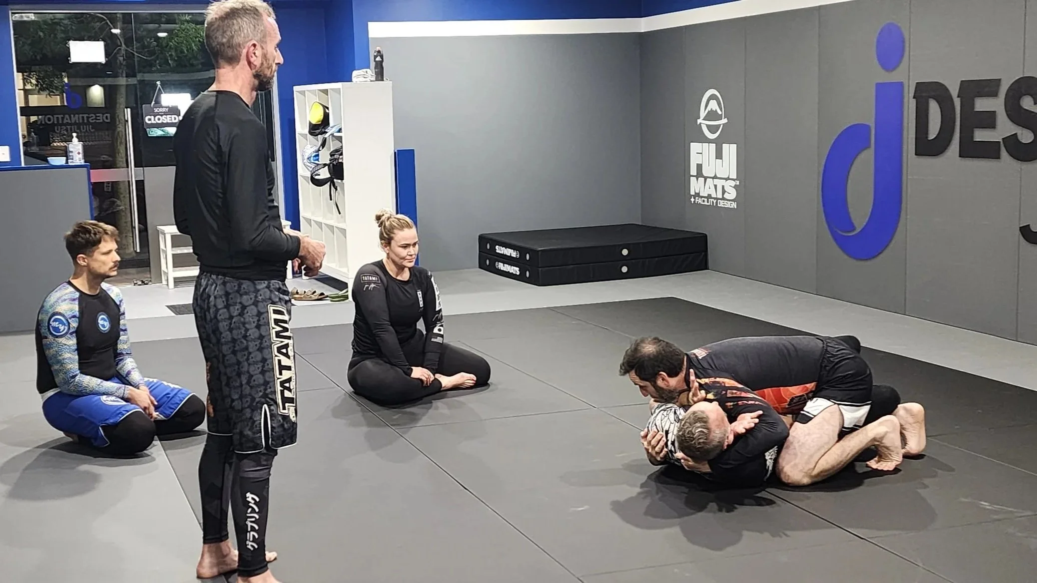 Three participants in a BJJ No Gi class watch a demonstration of a technique by a coach and an assistant. 