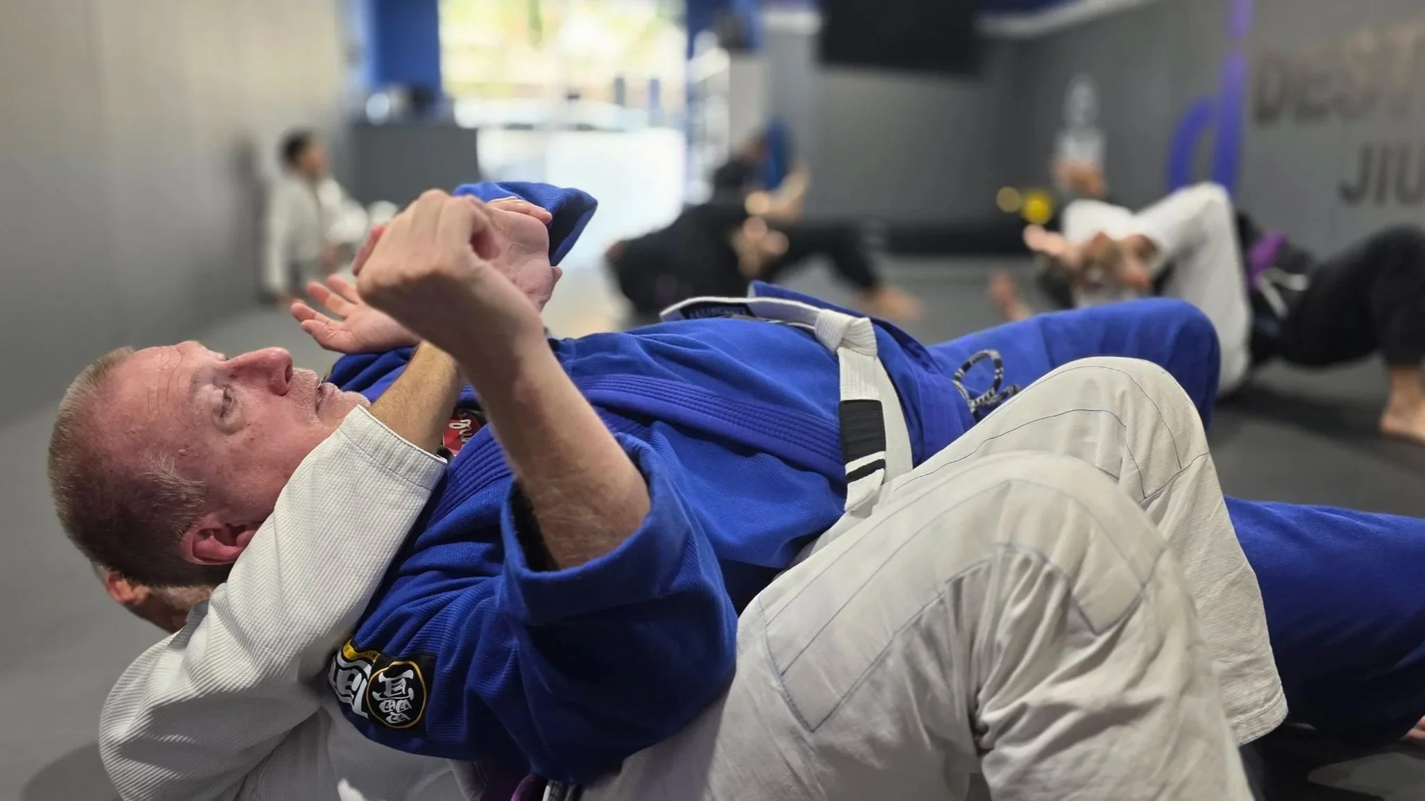A male student in a blue gi is defending against an attack from another male student in a white gi. Both students are on the ground, with the student in the white gi underneath the student in the blue gi. Other students are training on the ground in 