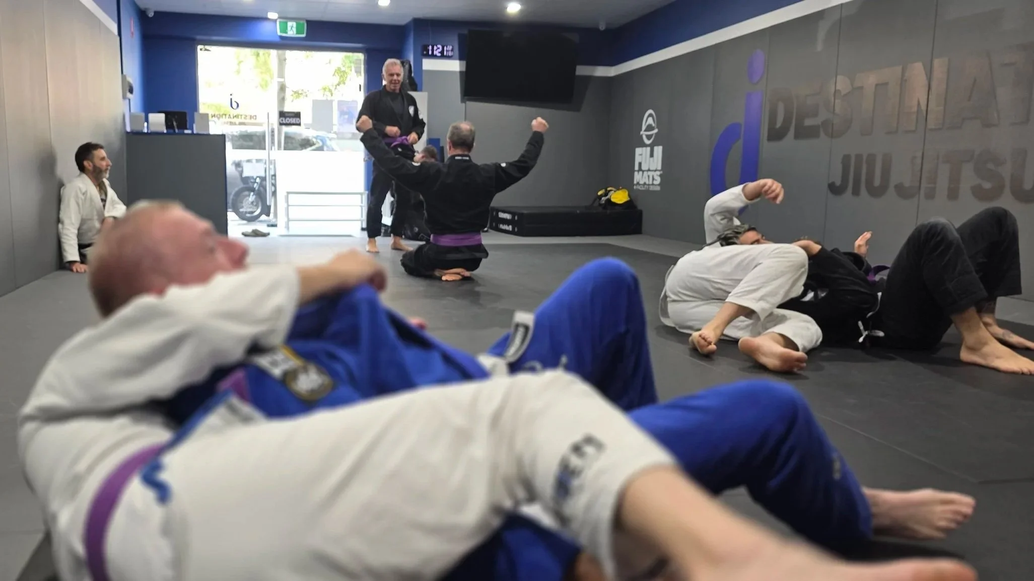 A group of students are training in the gi. There are three groups of two students. Two groups are actively practicing jiu jitsu moves. One student is holding his arms up, while kneeling on the gorund; his partner is standing, facing him. The coach i