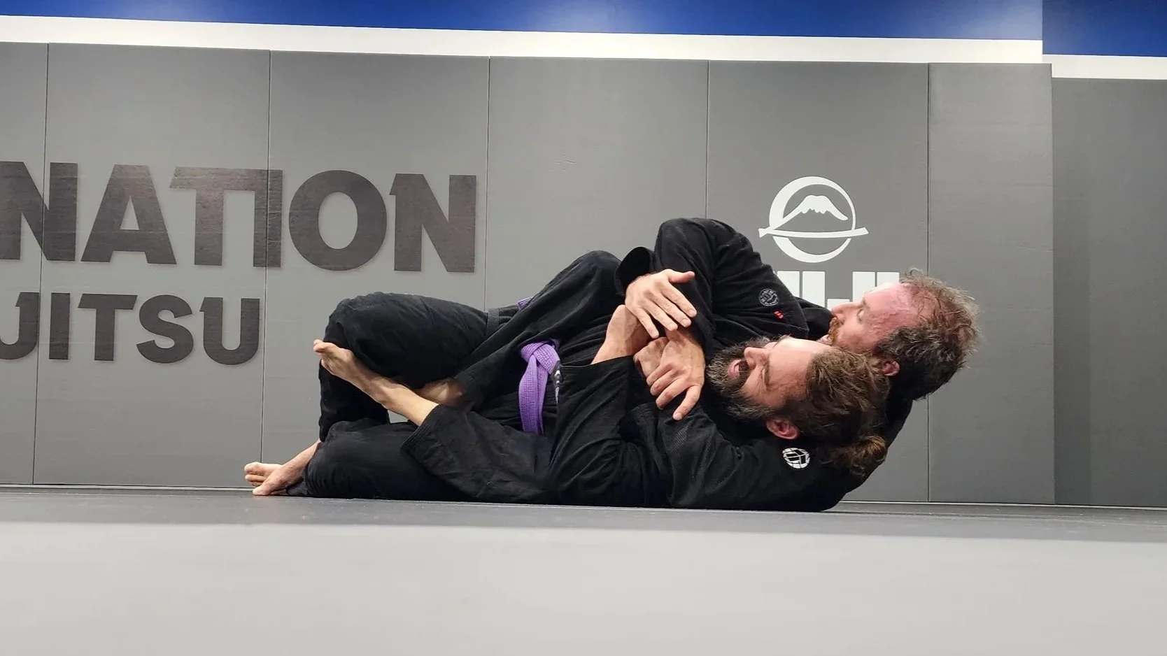 Two male students are practicing a strangle move while lying on grey mats. Boths students are wearing black gis, and one is wearing a purple belt, showing his rank. 