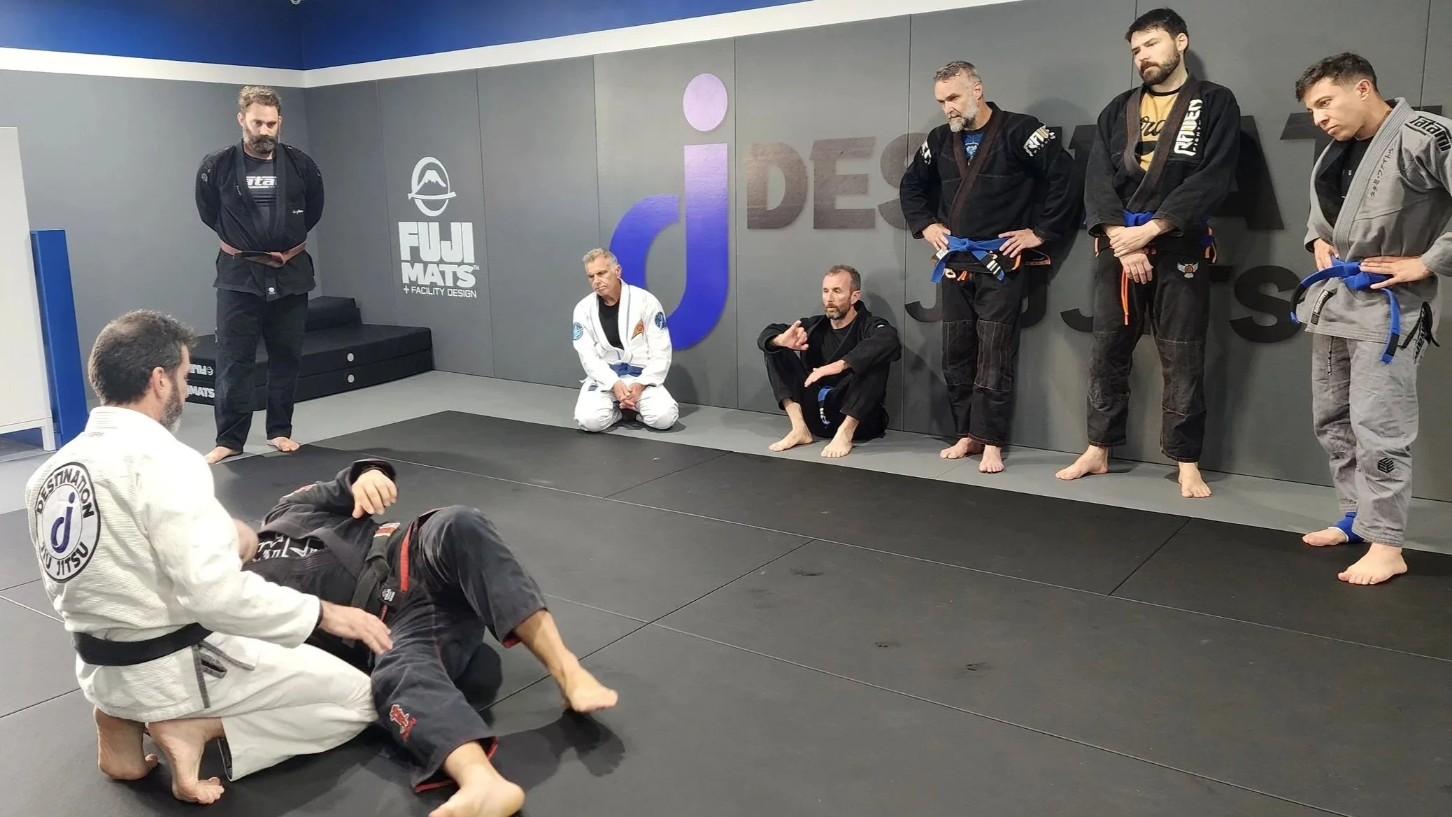 Six male students are watching a jiu jitsu technique demonstration. Four of the students watching are standing, and two are kneeling. The coach is wearing a white gi and is kneeling behind another student who is laying on his back. Everyone in the ro