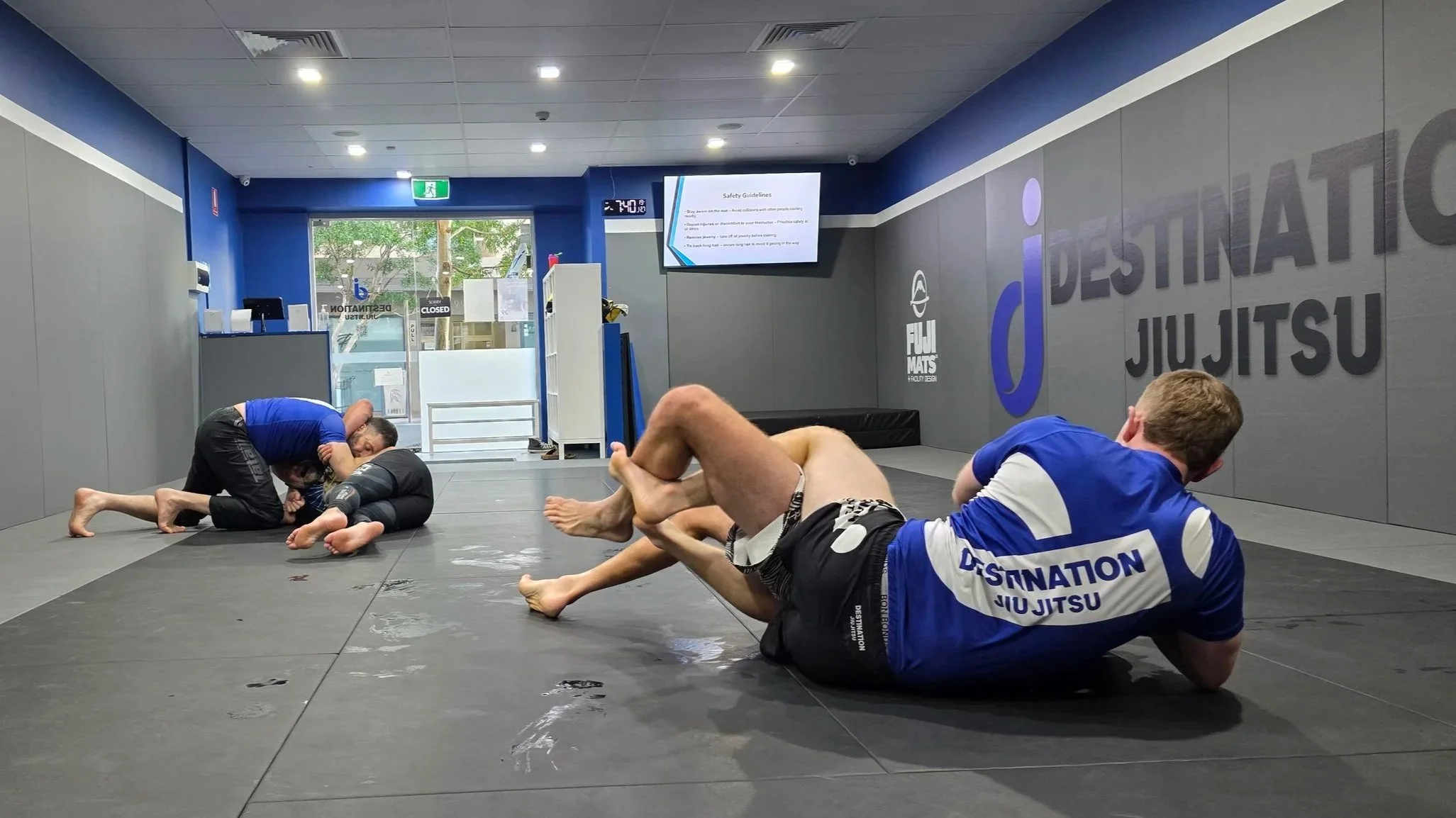 Two pairs of students are practicing no-gi jiu jitsu. The pair in the foreground have their legs tangled, with one student on his shoulder and his torso turned towards the camera. In the background, one student is lying on his side, with his partner 