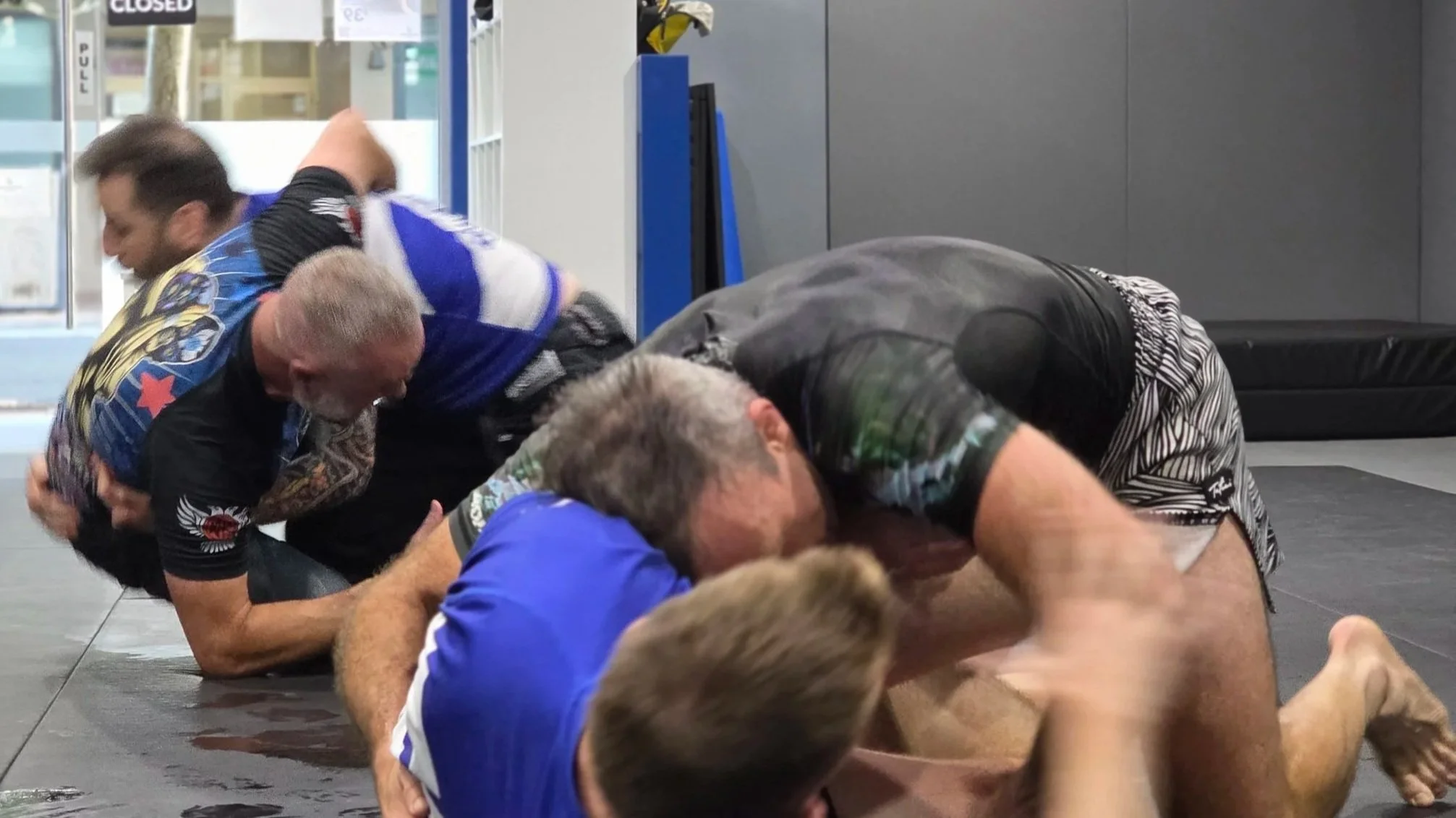 Two pairs of male students are actively sparring. They are all wearing short sleeve rash guards and grappling shorts. All four students are on the floor, grappling. 