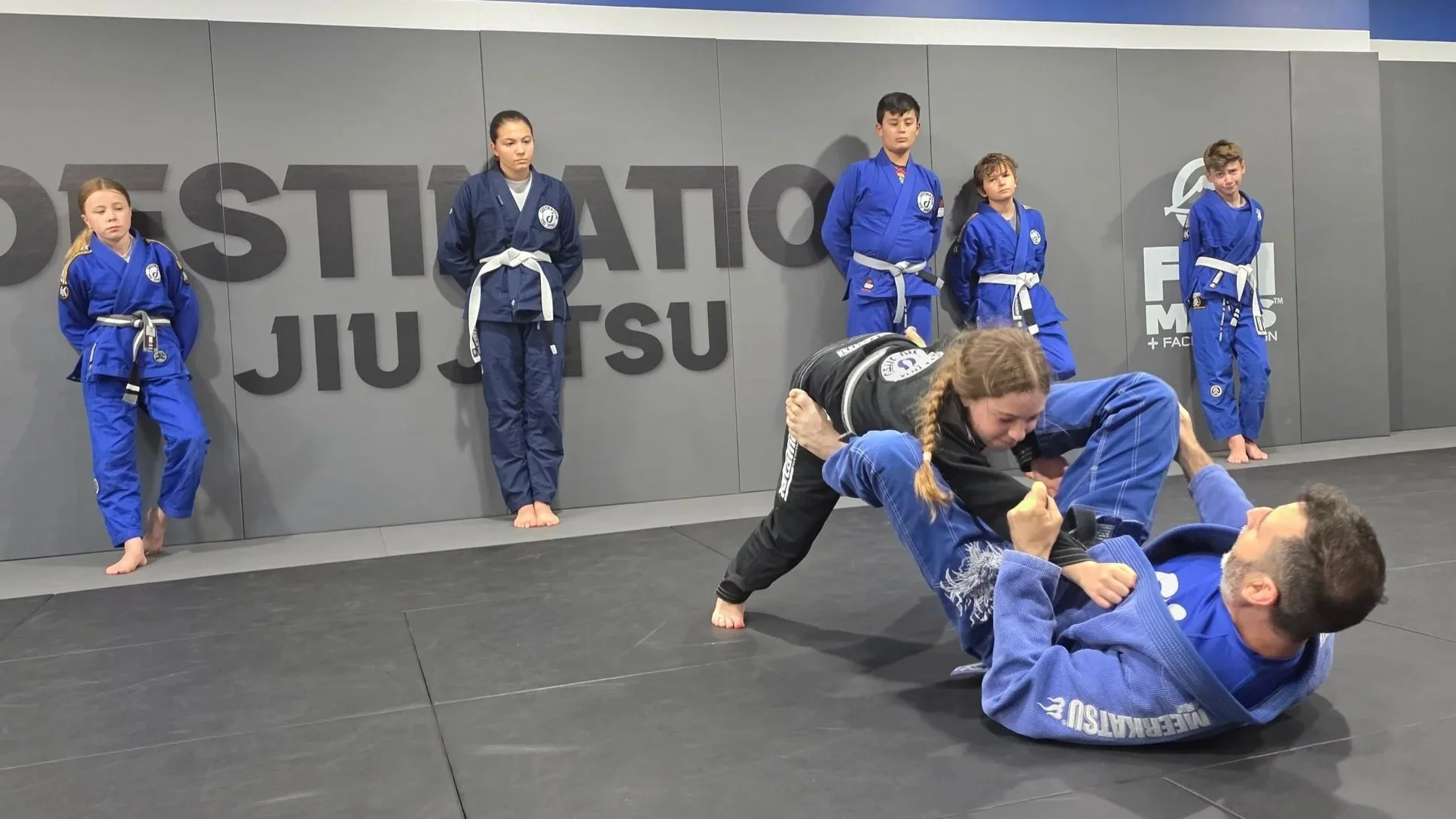 Five students in blue and navy gis are standing while they watch the coach demonstrate a technique with a female student in a black gi. The coach is lying on his pack with his hands holding the sleeves of the student in the black gi, and his legs are