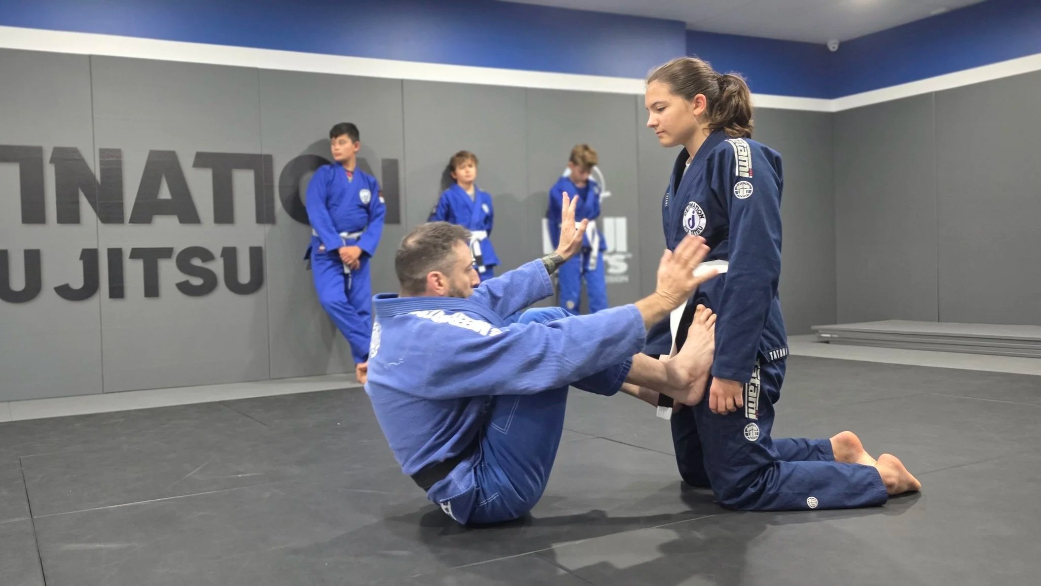 The coach is balancing on his bum with his legs partially bent. His feet are pressed against the thighs of the female student kneeling opposite him. The coach has his arms up and his palms flexed. There are three teenage male students wearing blue gi