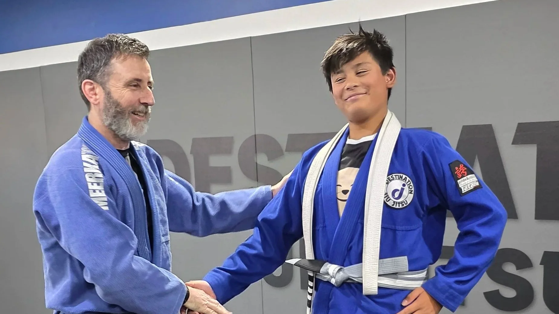 The coach is shaking the hand of a teens class student, a young male. Both are wearing blue gis. The teens class student looks happy as he was promoted to the next belt level. He has a white belt slung over his shoulders and a grey and white striped 