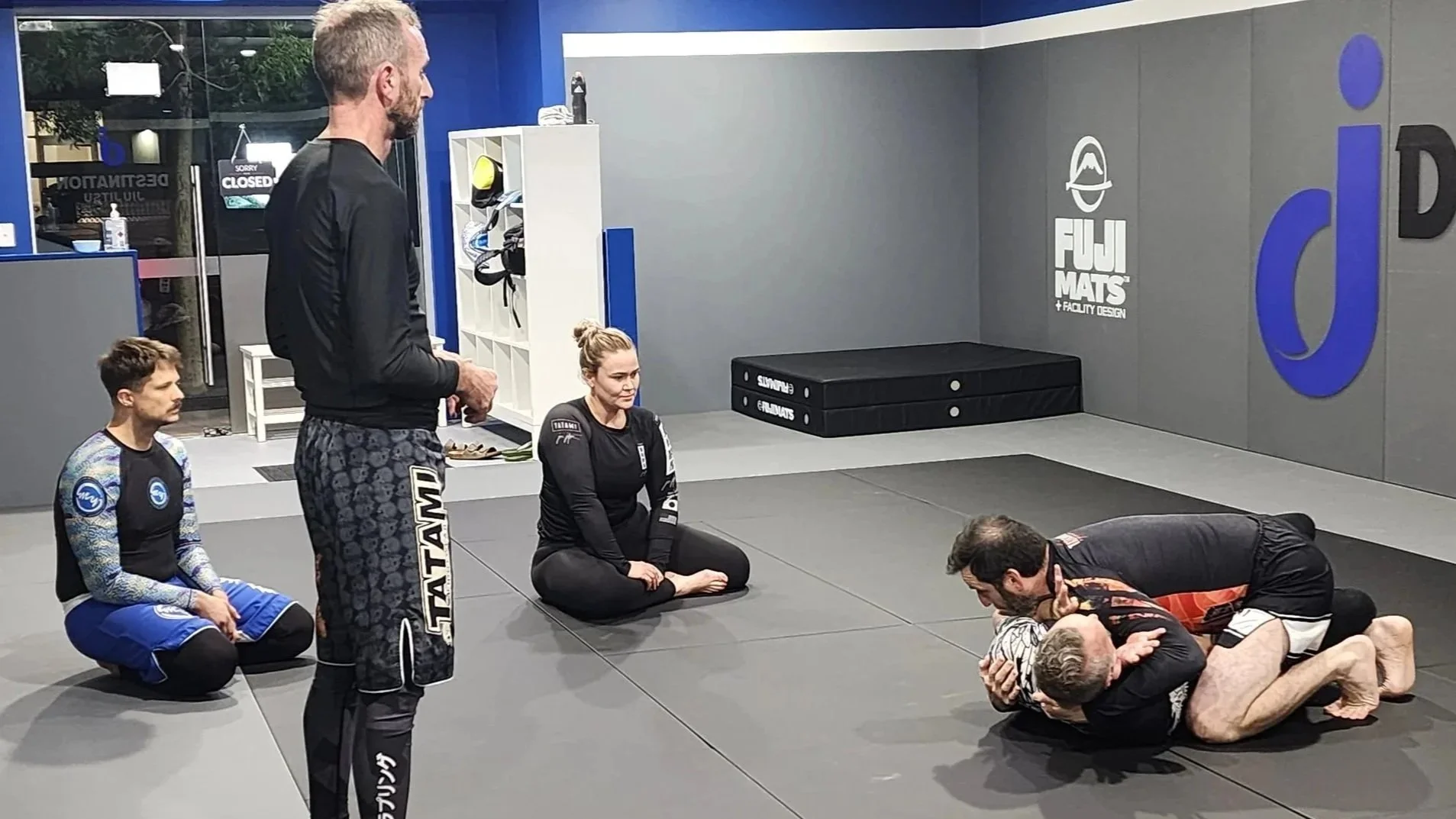 Three participants in a BJJ No Gi class watch a demonstration of a technique by a coach and an assistant. 