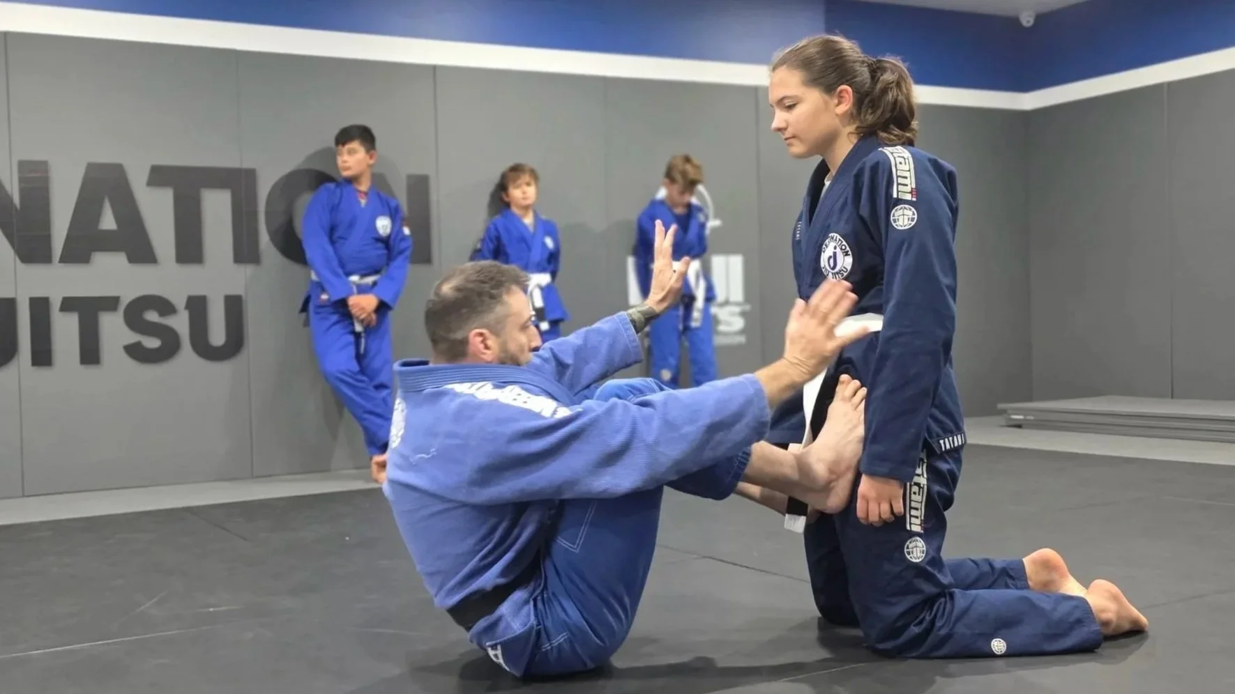 The coach is balancing on his bum with his legs partially bent. His feet are pressed against the thighs of the female student kneeling opposite him. The coach has his arms up and his palms flexed. There are three teenage male students wearing blue gi