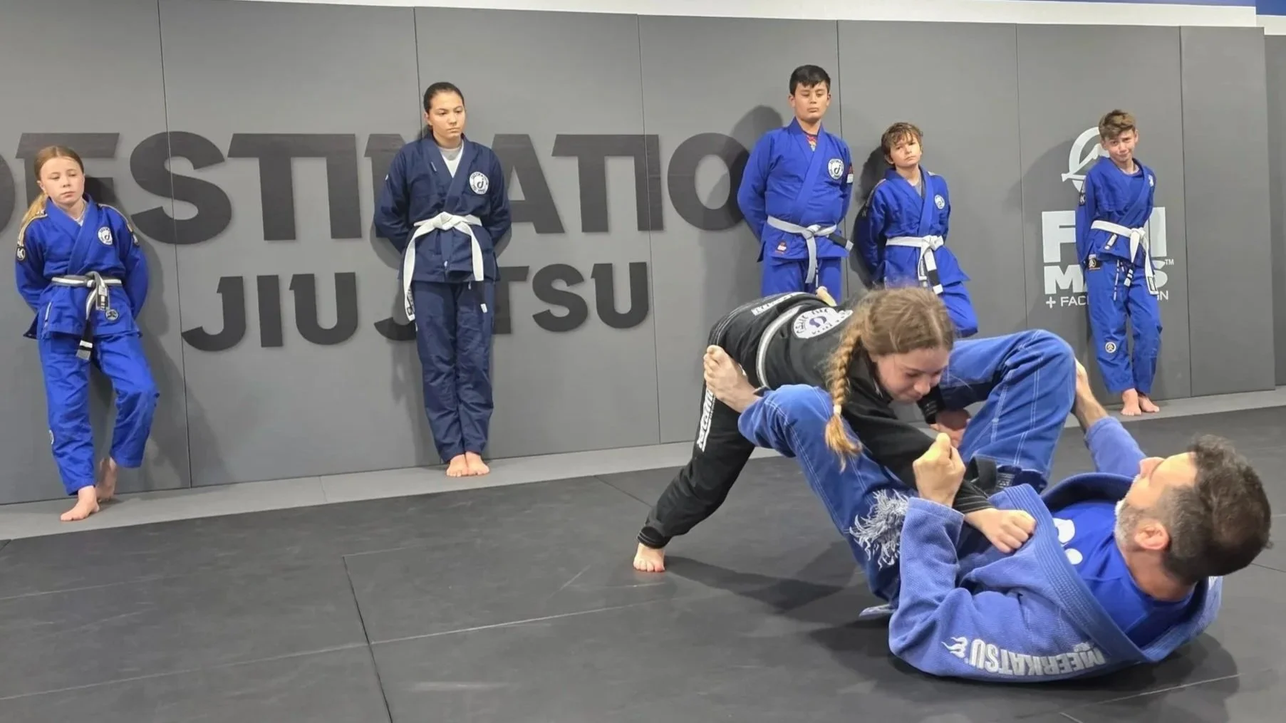 Five students in blue and navy gis are standing while they watch the coach demonstrate a technique with a female student in a black gi. The coach is lying on his pack with his hands holding the sleeves of the student in the black gi, and his legs are