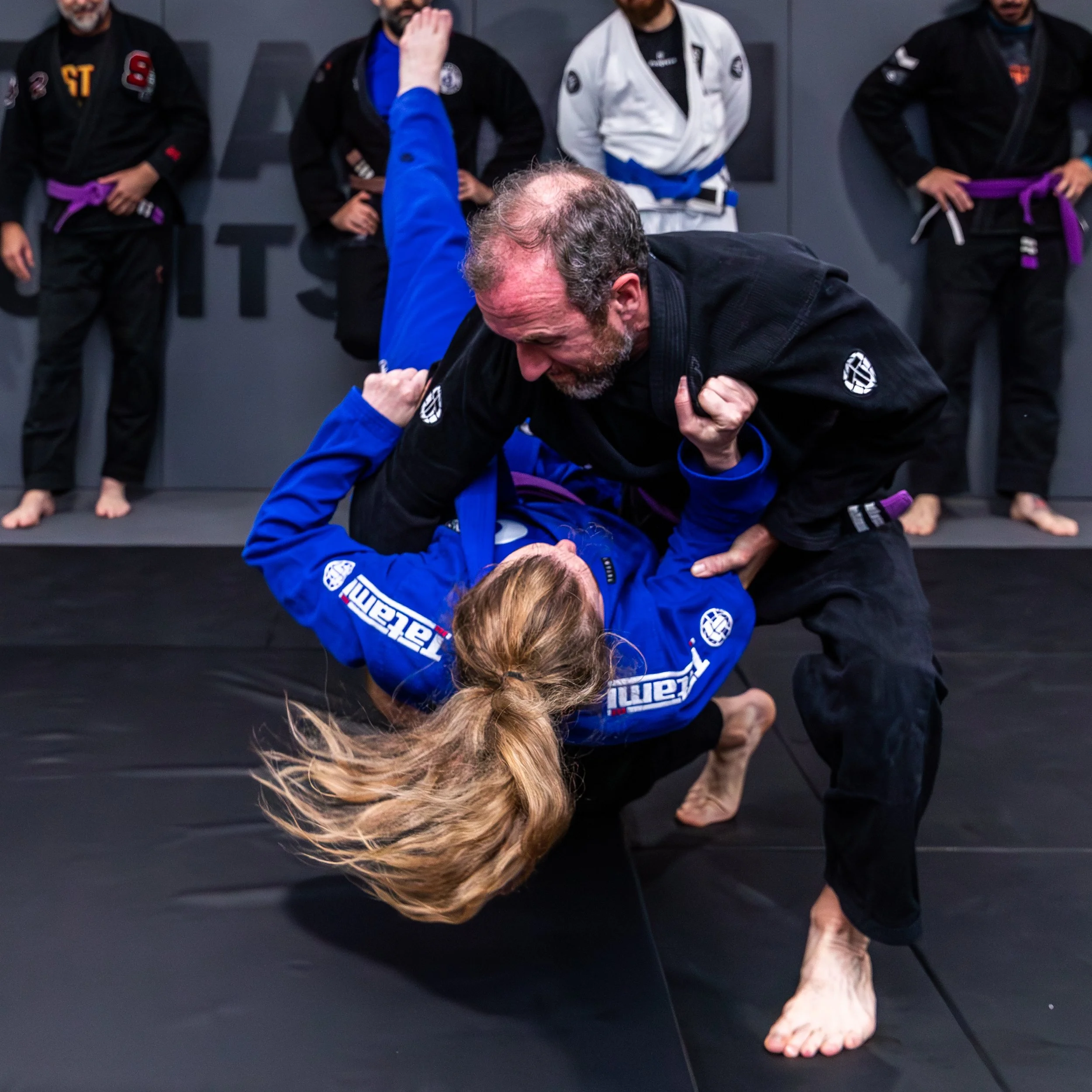 A male student is demonstrating a takedown throw of a female student. The Male student is wearing a black gi, and the female has long blonde hair and is wearing a bright blue gi. She is about to land on a black crashpad.