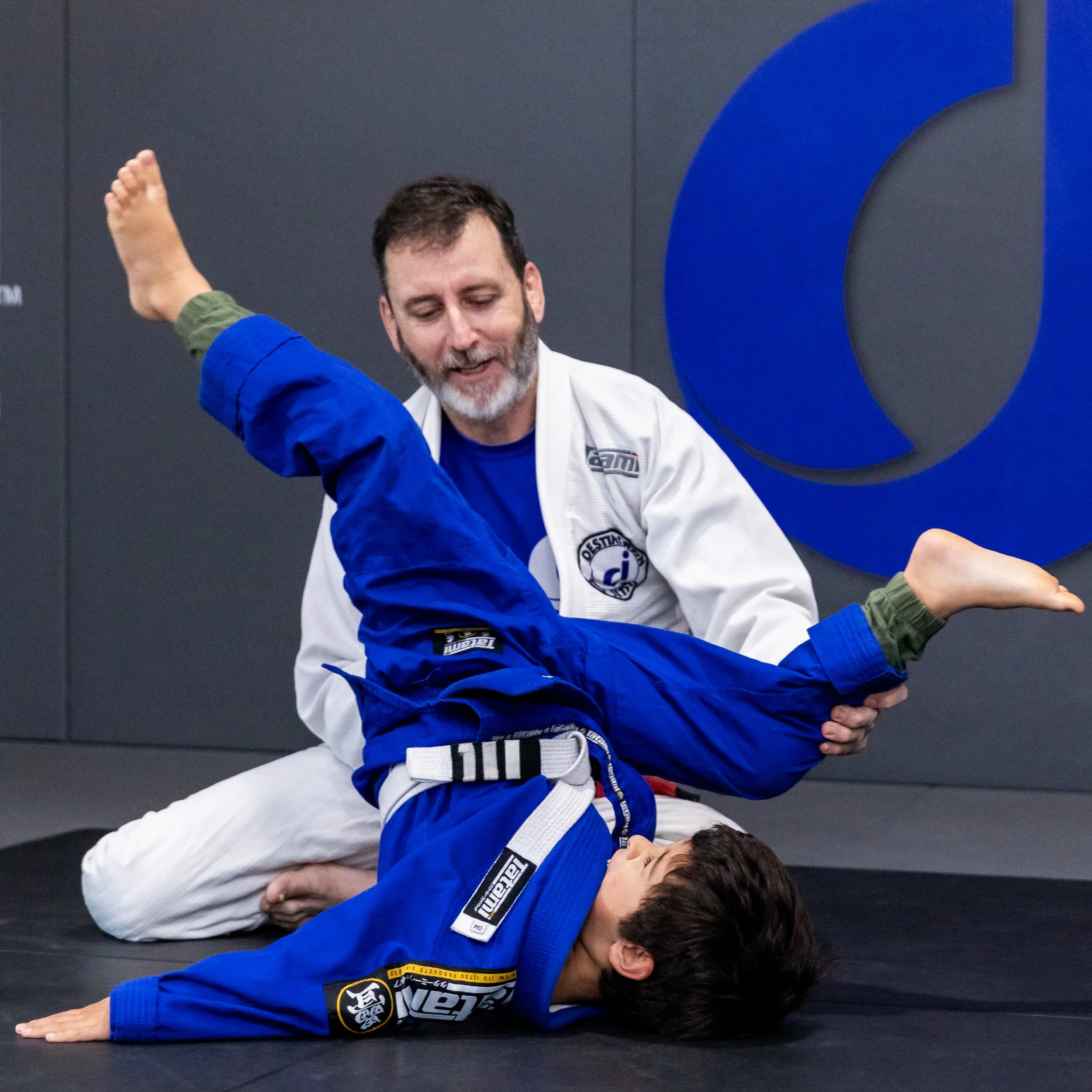 The coach, Trent, is helping a student to complete a backward roll. Trent is wearing a white gi, and has his hand on the student's knee. The student is upside down, with his shoulder and arm on the mat, with his legs in the air. He is wearing a blue 