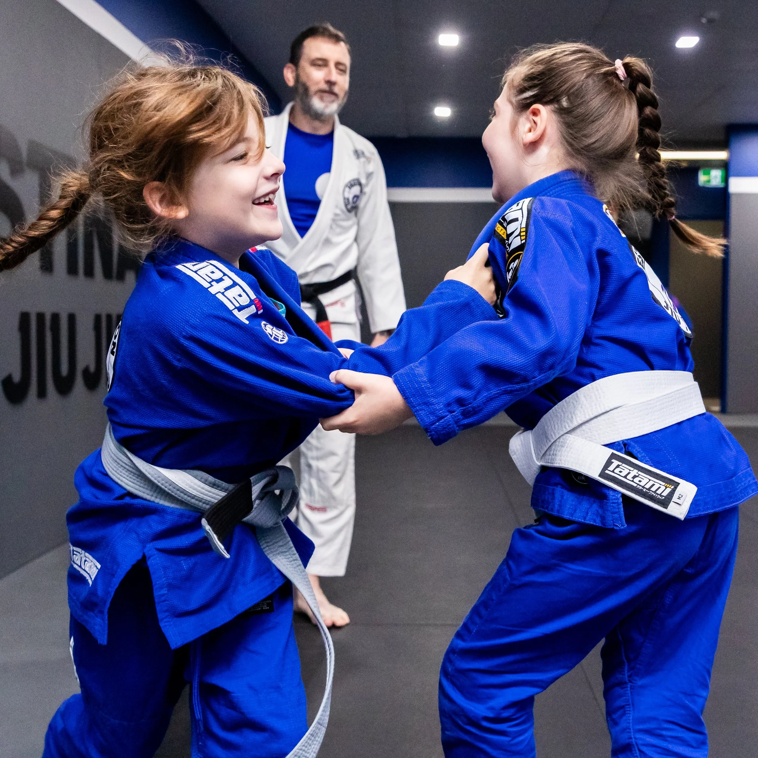 Two female students are playing a grips drill game. They are both wearing bright blue gis and have plaited hair. In the background, Coach Trent is watching the drills. He is wearing a white gi over a blue rashguard. Everyone is smiling.