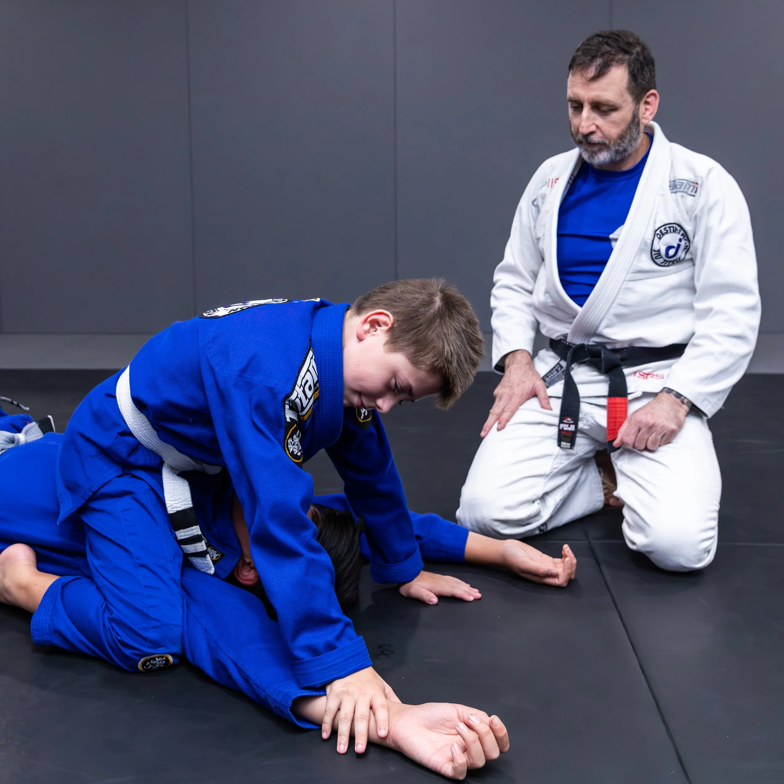 One teenage student is sitting on the chest of another, with his knees bent and shins on the floor. One of his hands is pinning the wrist of the other student, and his other is resting on the mat. Both students are wearing blue gis. The coach is watc