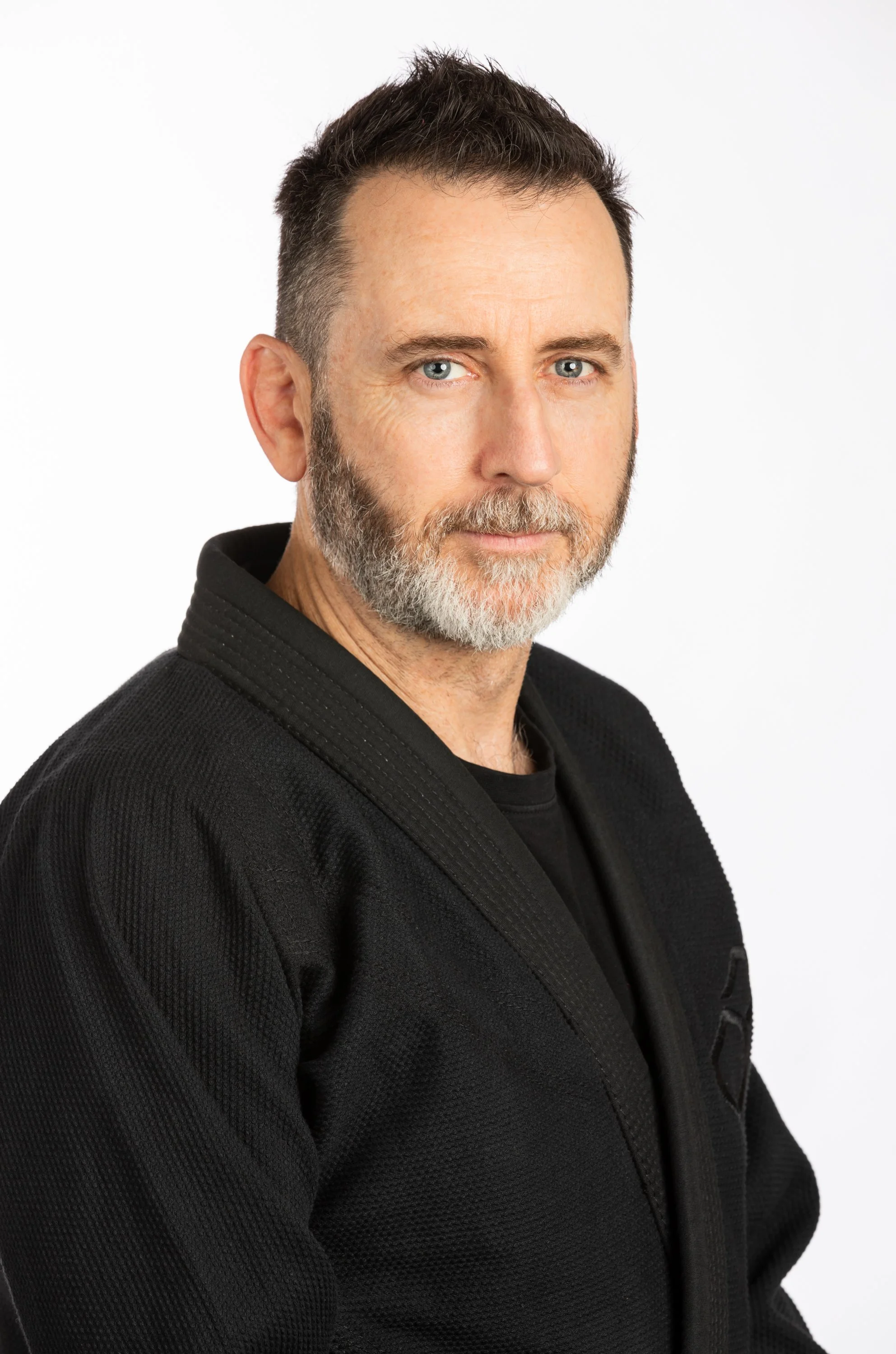 Image of Trent, the head coach. Trent is wearing a black gi jacket over a black rashguard. He has short, dark hair, styled to a slight point. He has a short grey and dark brown beard. He has a slight smile on his face, and is facing the camera.