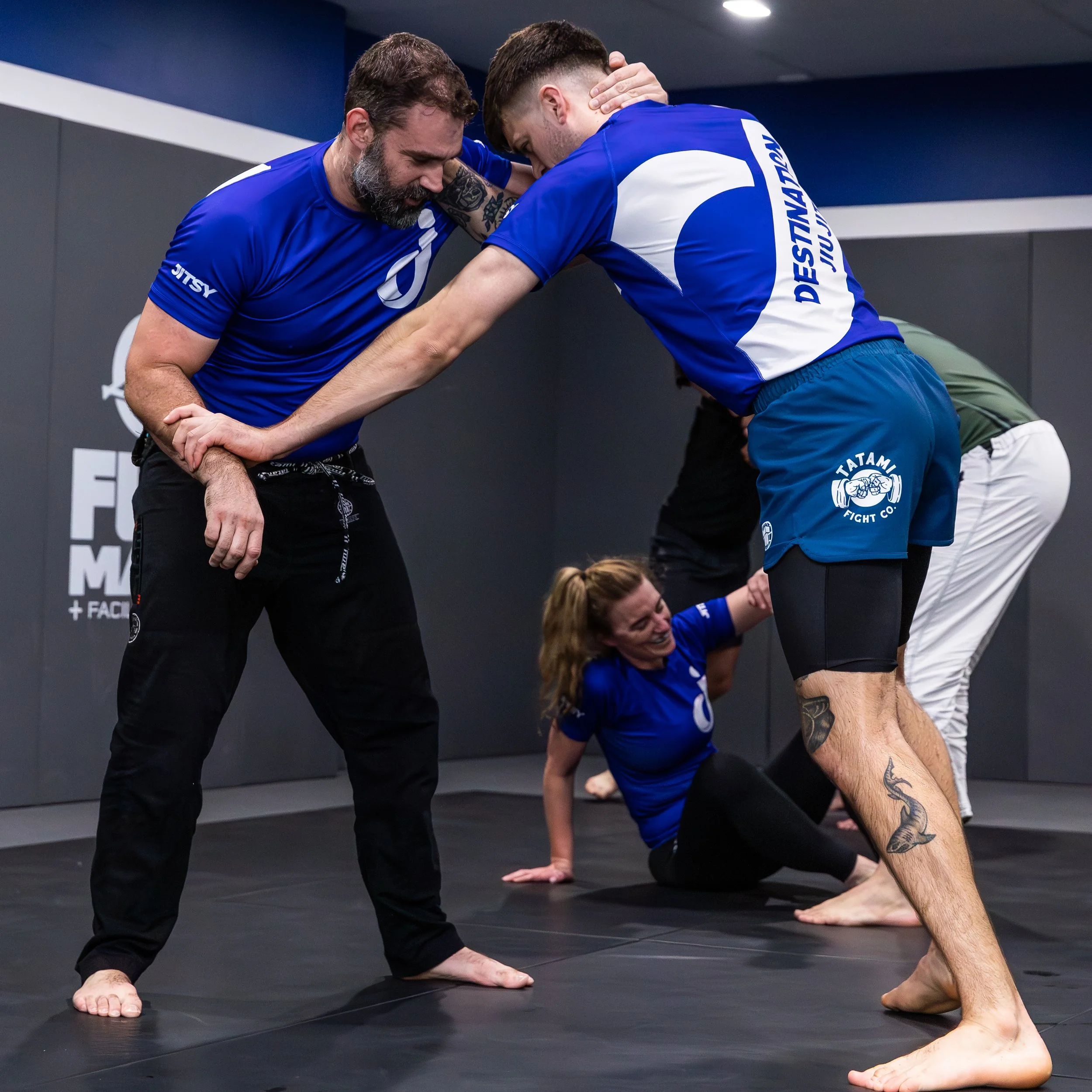 Two adult students are standing, beginning to grapple. The student in front is holding the wrist of his opponent with his left hand, and has his right hand on the back of his opponents neck. The other student has his hand cupped around the back of th