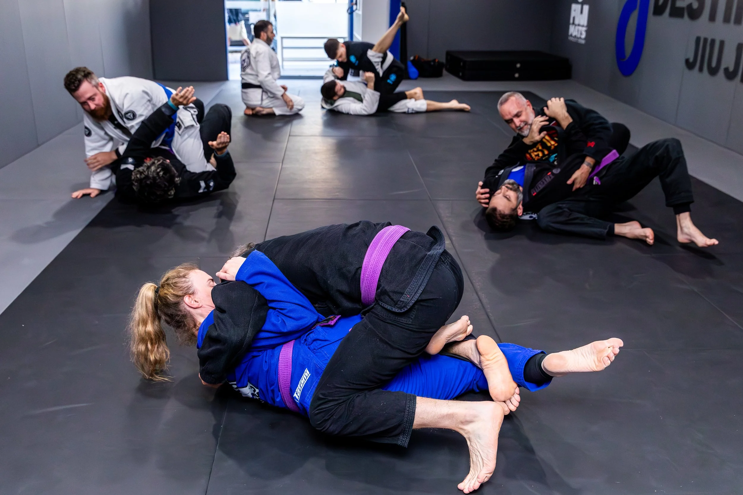 Four pairs of students in gis grappling on black mats. The coach, Trent, is on his knees next to one pair of students in the background, coaching.