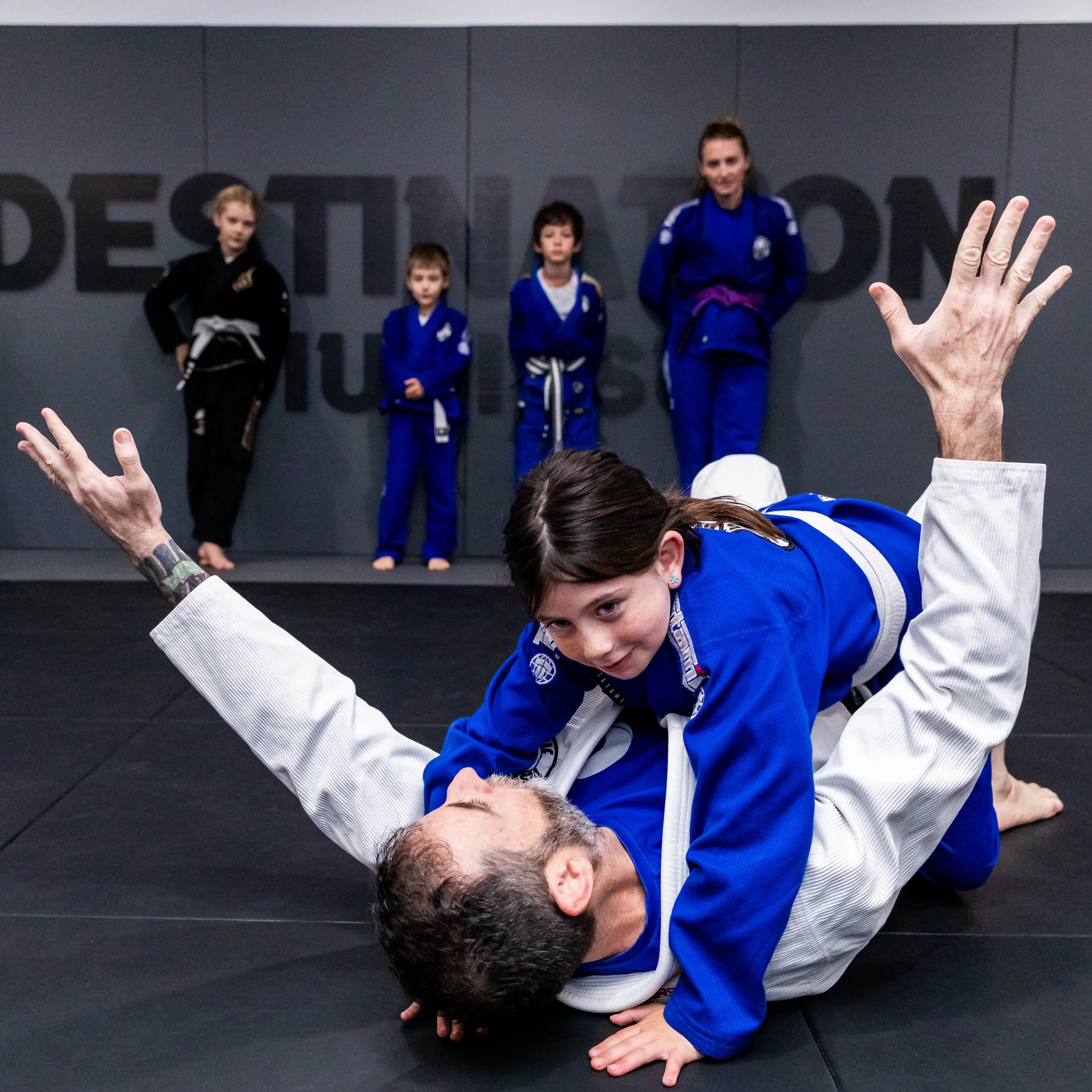 A female student is demonstrating a pin technique on the coach, Trent. The student has her chest on the coaches, with her arms over his shoulders. She is wearing a blue gi, and has black hair. Trent is laying on his back with his arms in the air and 