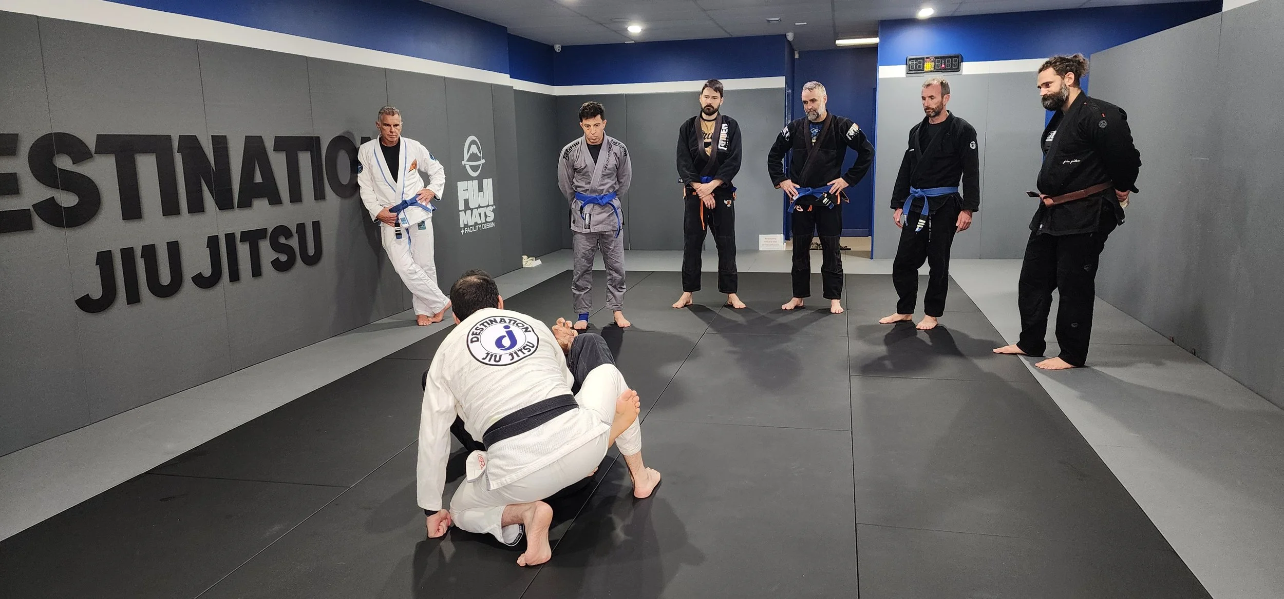 A group of adults in Brazilian Jiu Jitsu kimonos standing and watching a demonstration of a technique by 2 males in a white and black kimono. the floors and walls are covered in grey paded mats. The logo of the studio is printed on the pads.