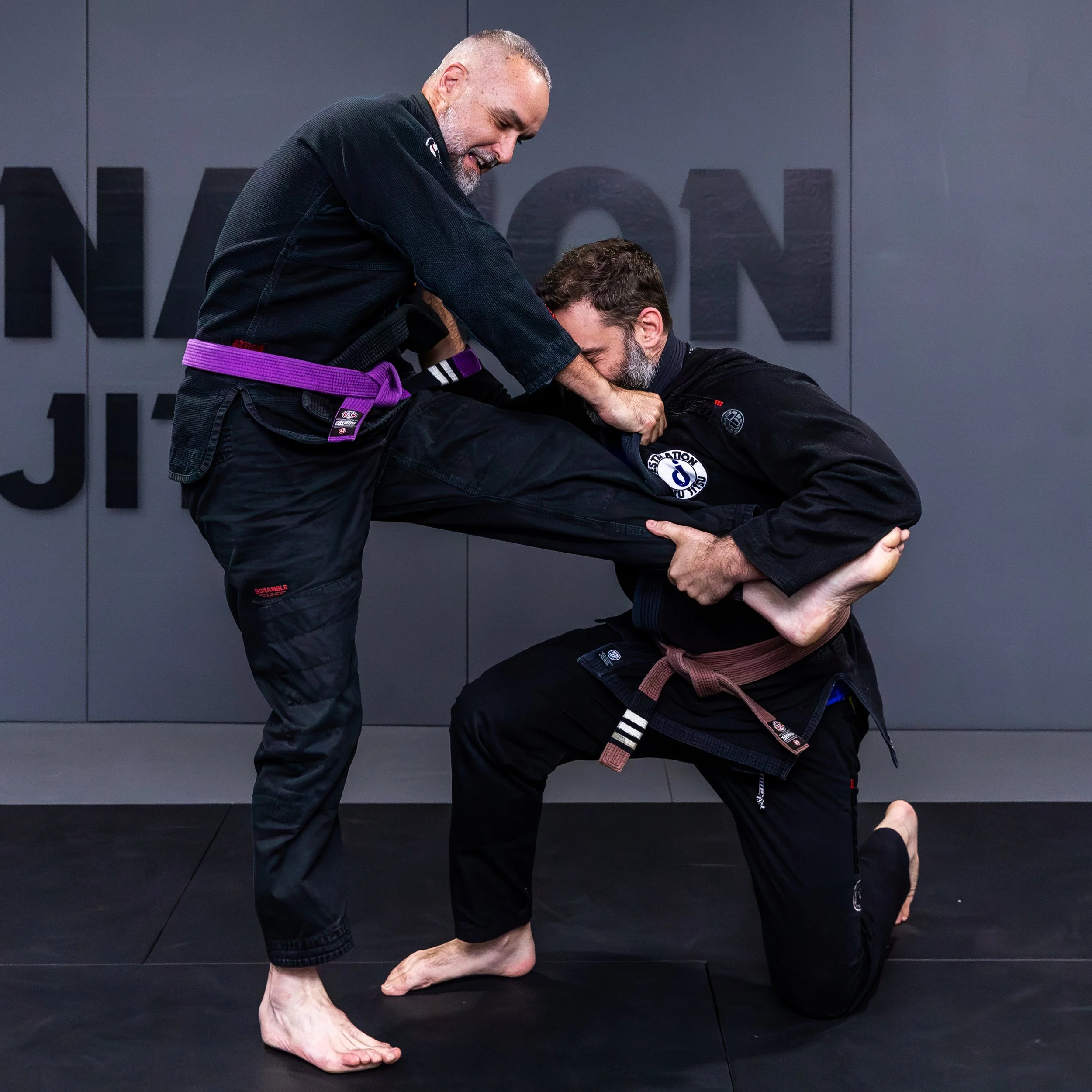 Two male students in black gis are practicing a single leg takedown move. The student wearing a brown belt is on one knee, gripping the other student's jacket with his right hand, and the right shin of his opponent with his left hand. The other stude