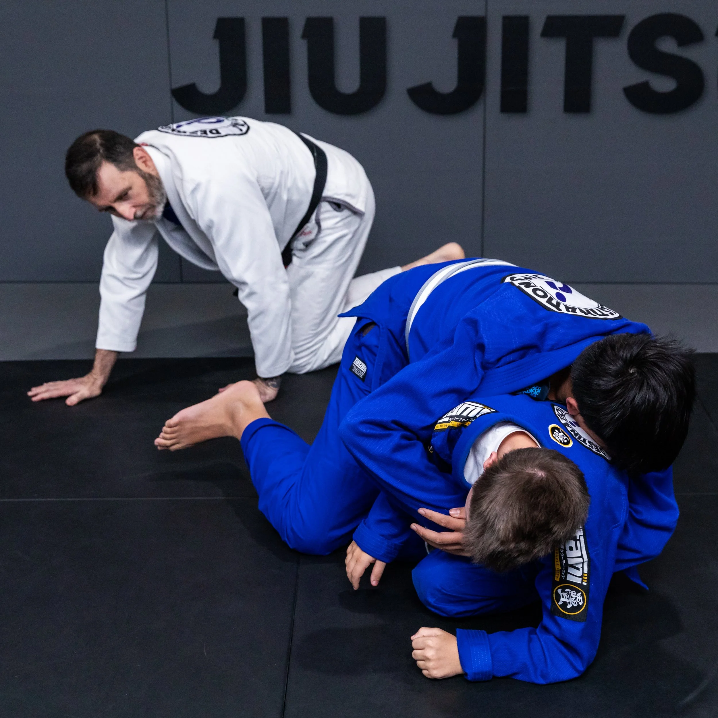 Two teen male students are in a scramble on the mats. Both students are wearing blue gis. In the background, coach Trent is on his hands and knees, watching the action. He is wearing a white gi.