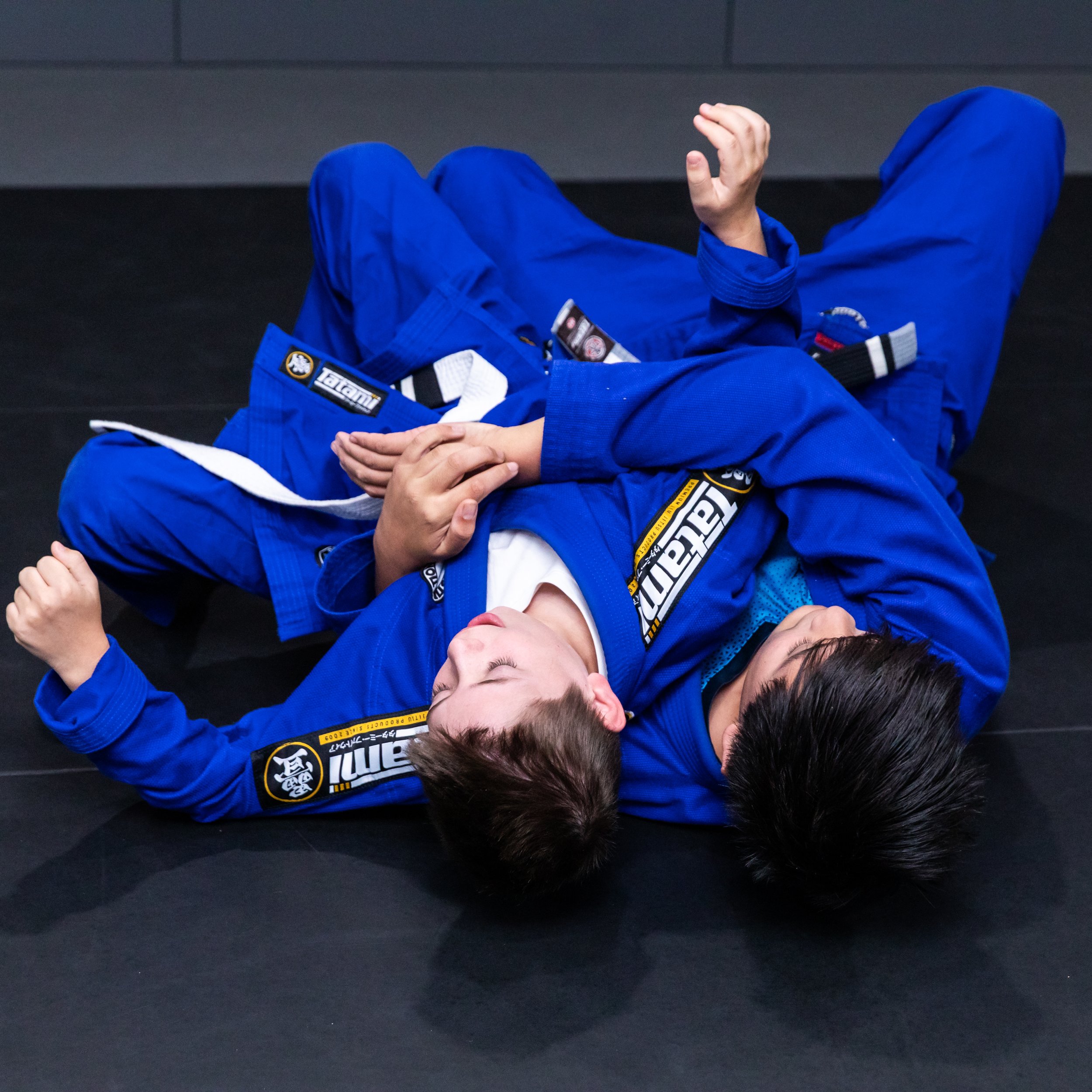 Two teenage students are laying on the mat with their heads towards the camera. One student has a seatbelt control with his arms around the other student. Both are wearing blue gis and have dark hair. 