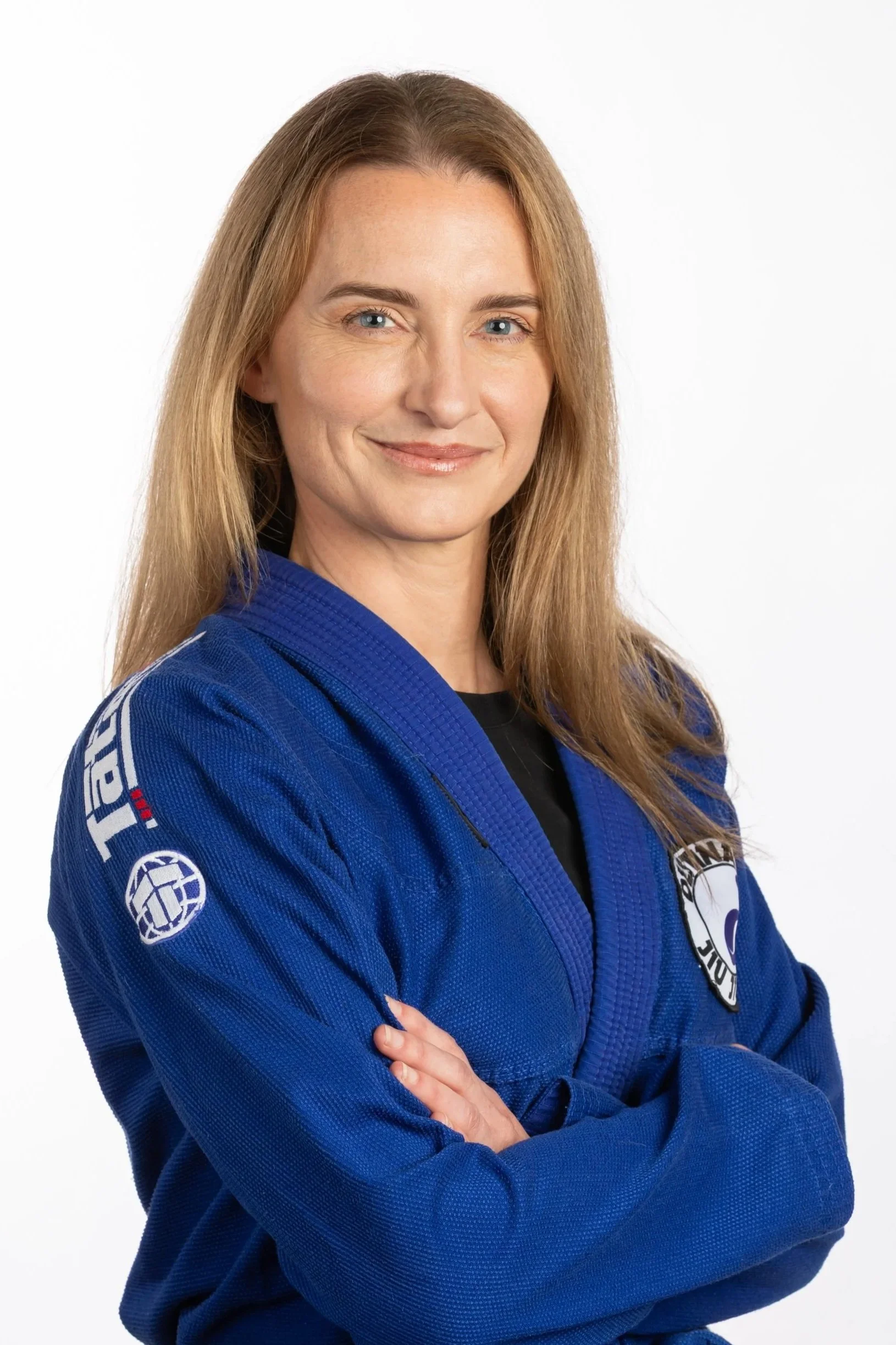 Karina is wearing a bright blue gi jacket over a black top. Her gi has a gym logo patch on the left lapel. Her arms are folded and she is facing slightly away from the camera. She has long blonde hair and a a closed mouth smile.