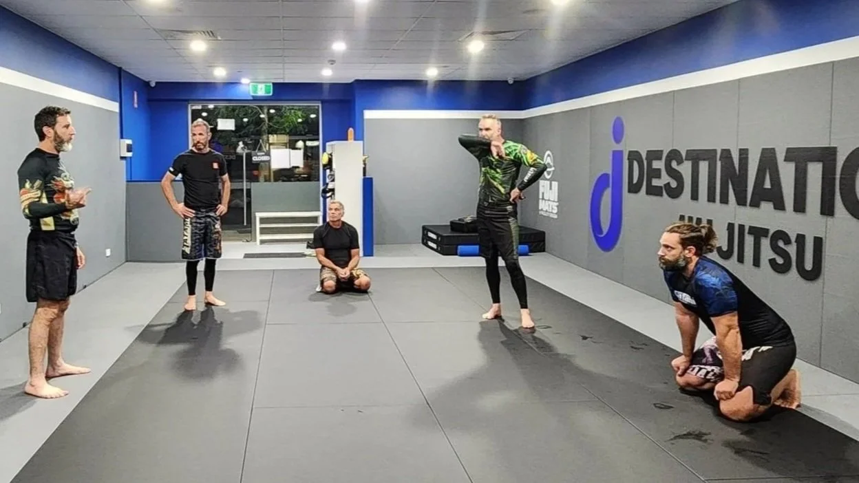 Four class participants listening to the coach explain a technique. Two participants are kneeling, and two are standing. All participants are wearing colourful rashguards and grappling shorts. 