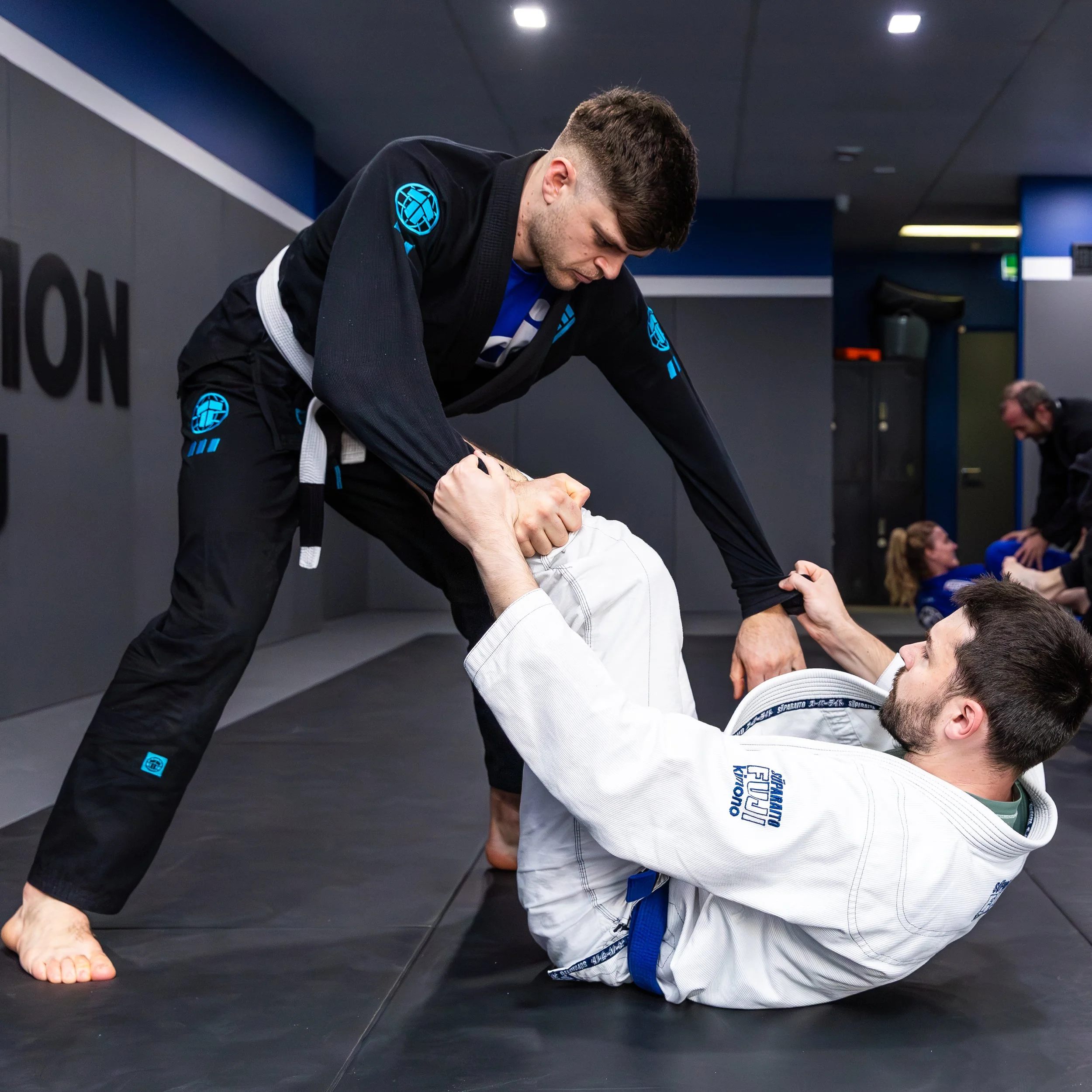 One student, a male in a white gi with a blue belt, is balancing on the ground on his lower back. He is holding the sleeves of his opponent, a male in a black gi with a white belt. The student in the white gi has his left eg extended, with his foot o