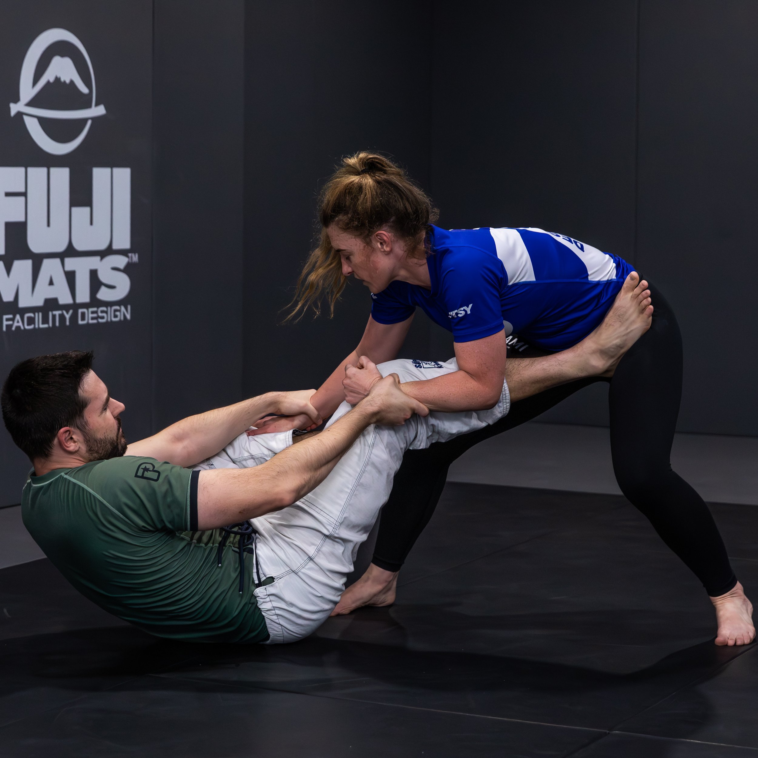 A female student with a blonde pony tail and a blue rashguard is standing and bent forwards at the waist, attempting to go to a mounted position on her opponent. The male is wearing a khaki short sleeved top and white pants. He is on his bottom, his 