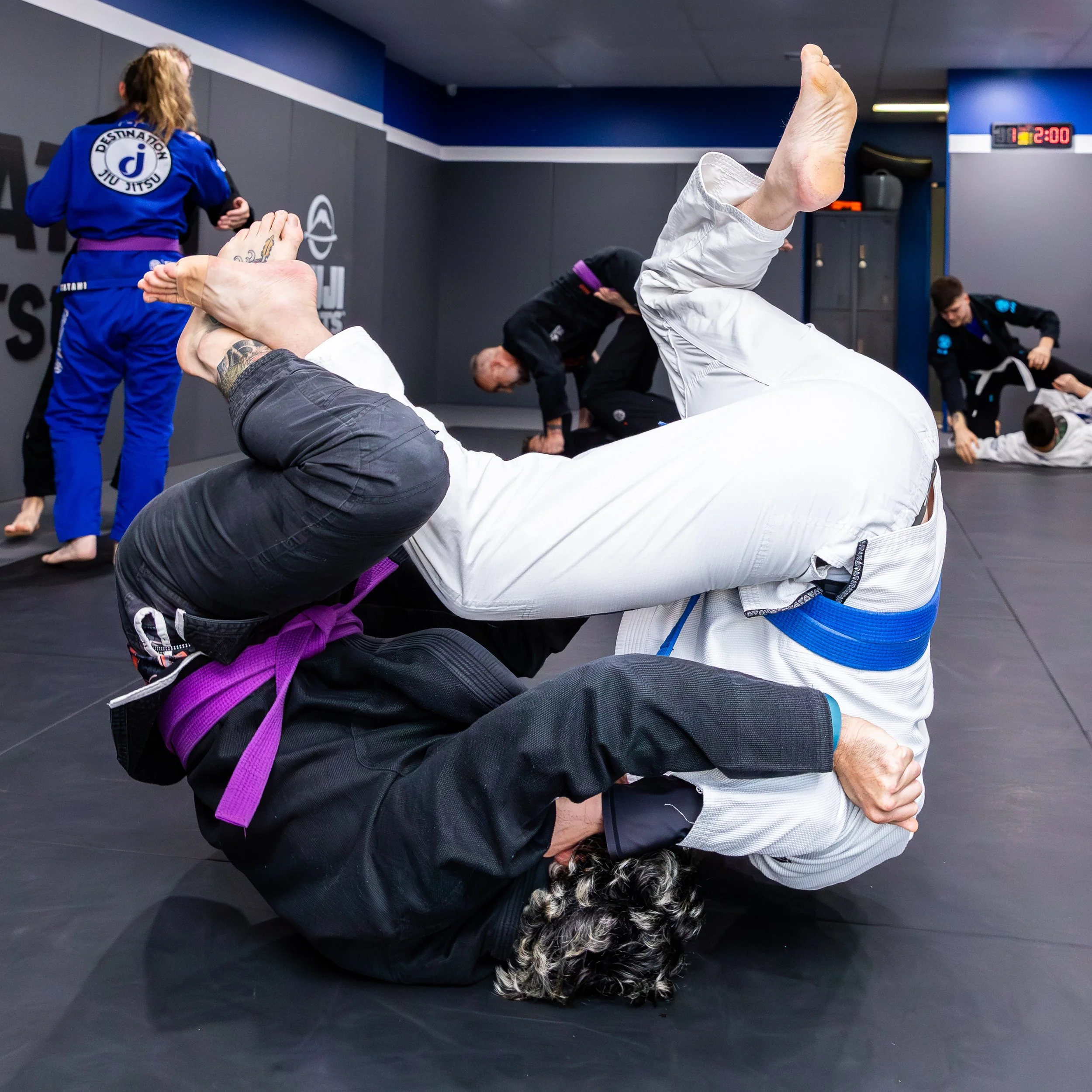 The image shows a busy gi class. Two students are in the forground wrestling. they are upside down, with their ankles touching, and their arms gripping each other. In the background are three pairs of students, in various positions. 