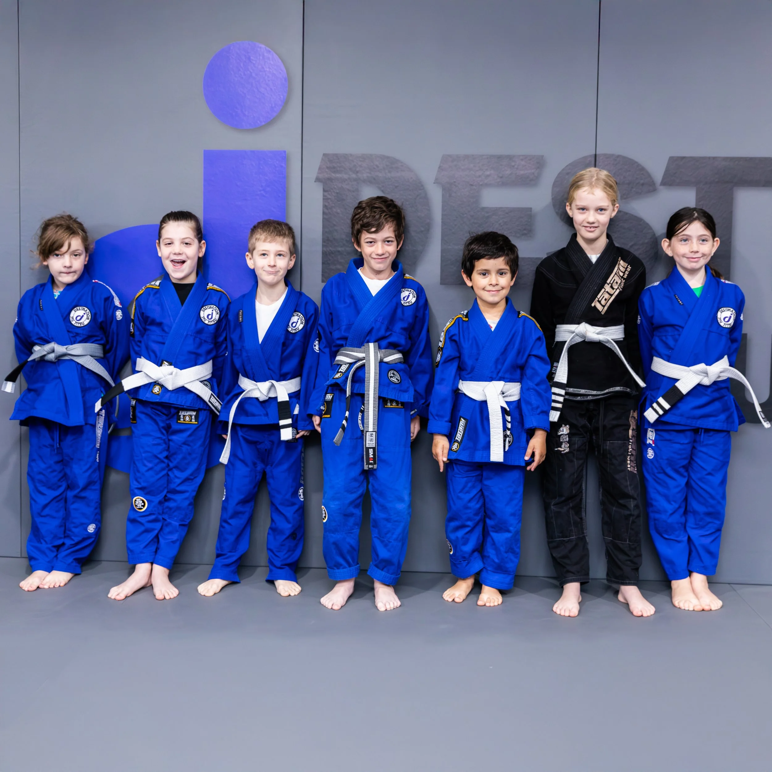 Seven chilren wearing blue and black gis standing against the grey padded wall. The children are smiling and facing the camera. The gym's logo is on the wall in the background.